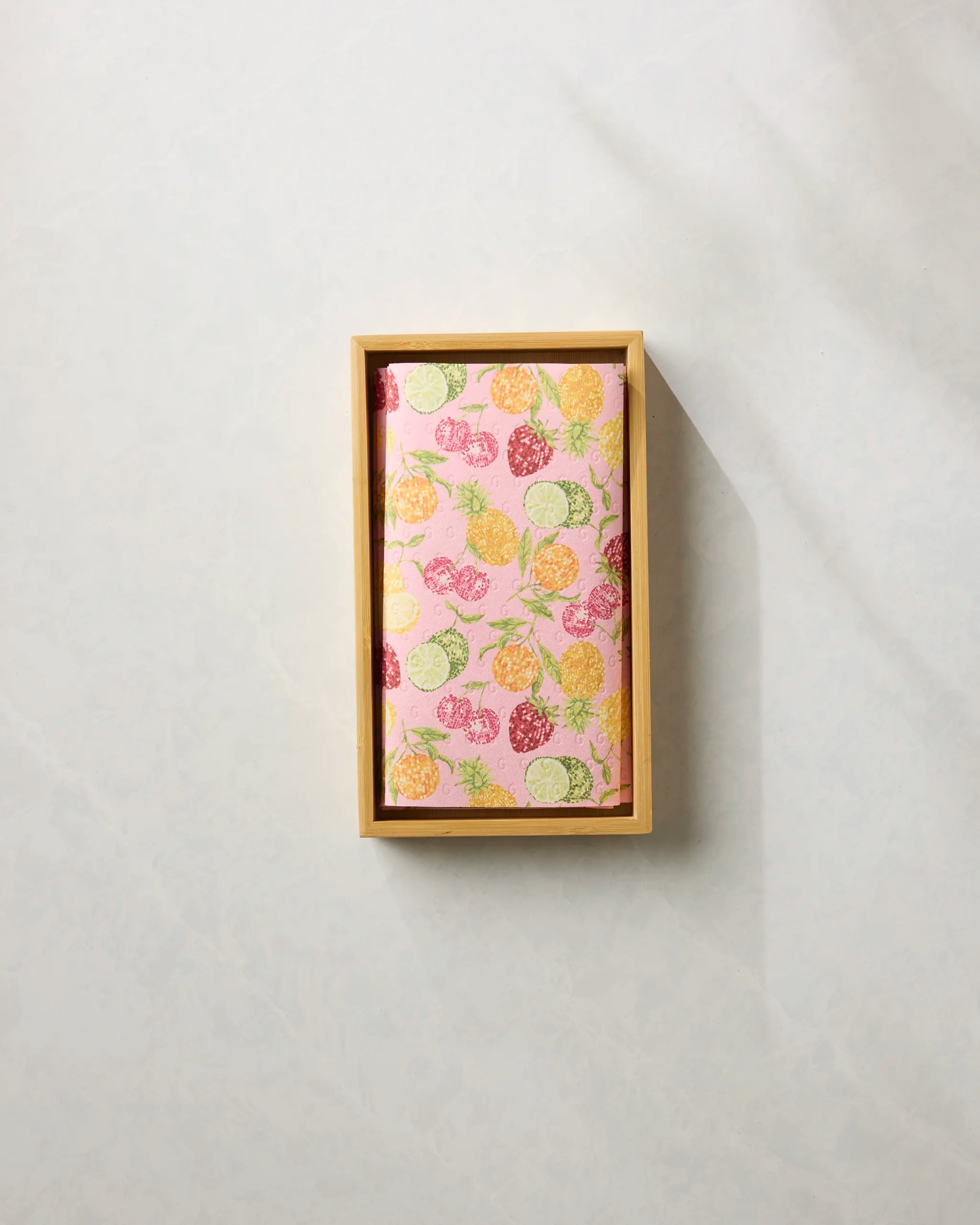 A rectangular wooden tray displays Geometry’s Disco Fruits pink notebook with colorful fruit designs and eco-friendly paper towels, set against a light background.