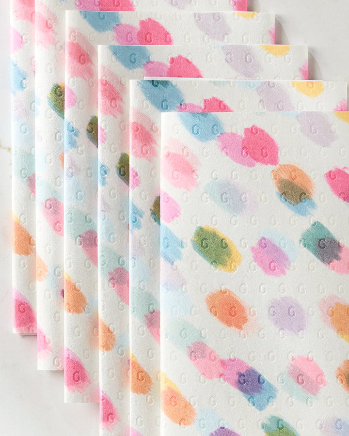 Six stacked, reusable Downpour towels by GEOMETRY feature pastel swatches in pink, blue, orange, green, and purple. Each white eco-friendly napkin is embossed all over with small repeating “G” letters.
