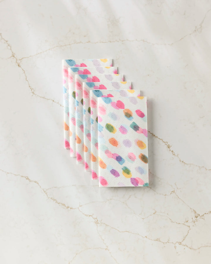 Six stacked GEOMETRY Downpour reusable paper towels in a pastel polka dot pattern are arranged neatly on white marble with beige veining.