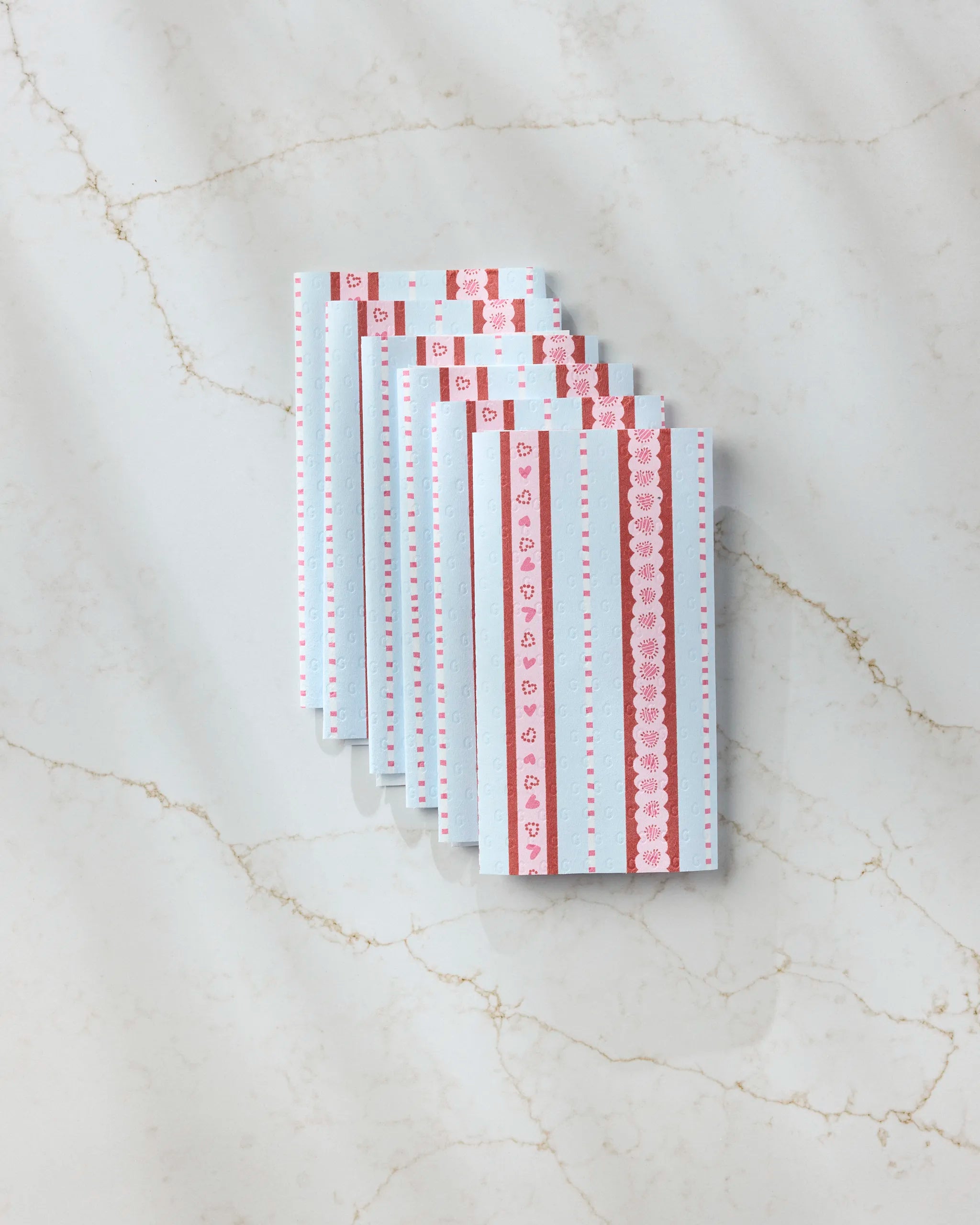 Geometry’s Dreaming of You envelopes, featuring blue and white heart and lace patterns, are fanned out on a marble surface next to Geometry’s eco-friendly paper towels for a charming, sustainable touch.