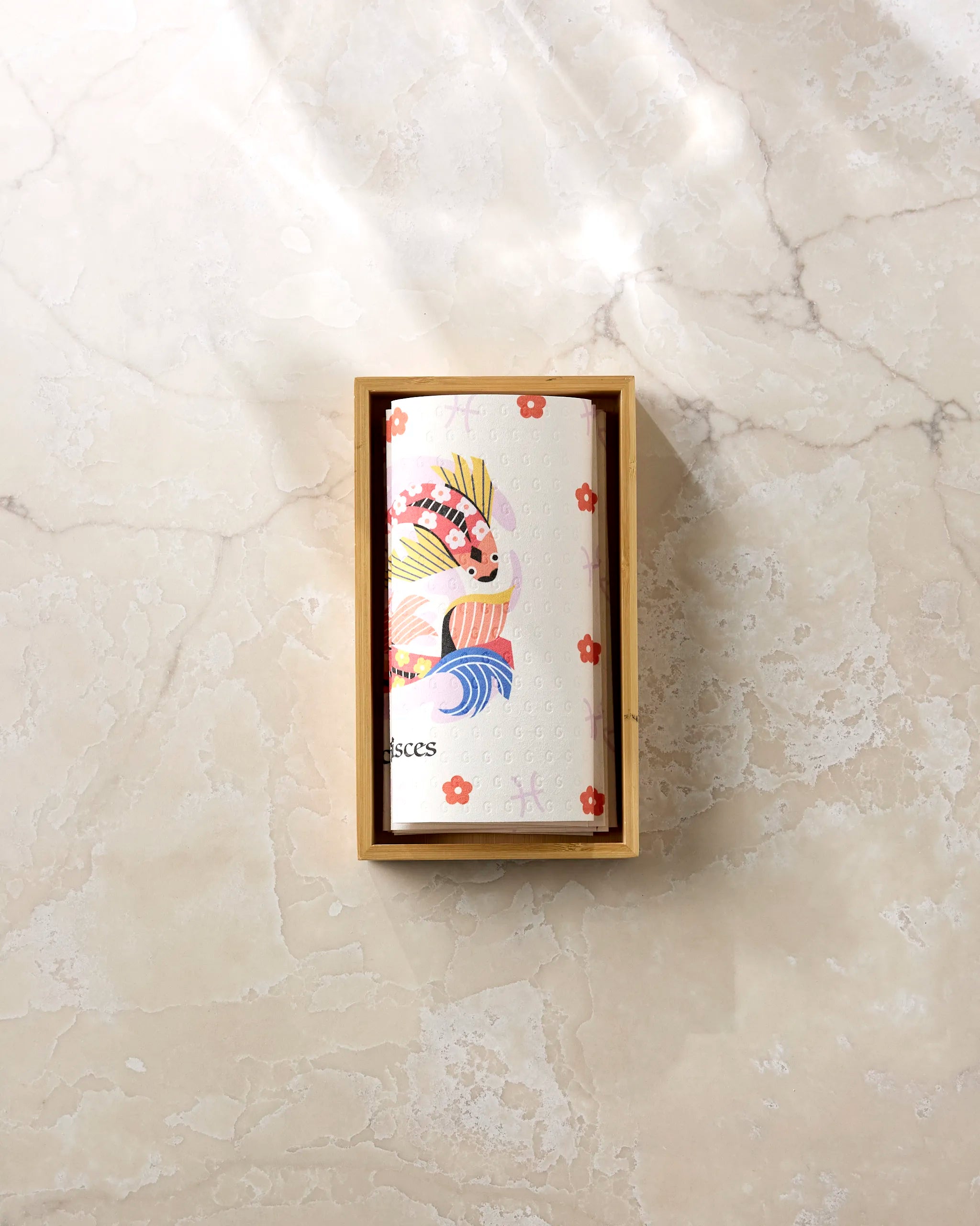 The Dreamy Pisces eco-friendly paper towels by Heather Zhou, featuring a colorful fish design, are displayed in a wooden box on a light beige marbled surface with sunlight casting soft shadows.