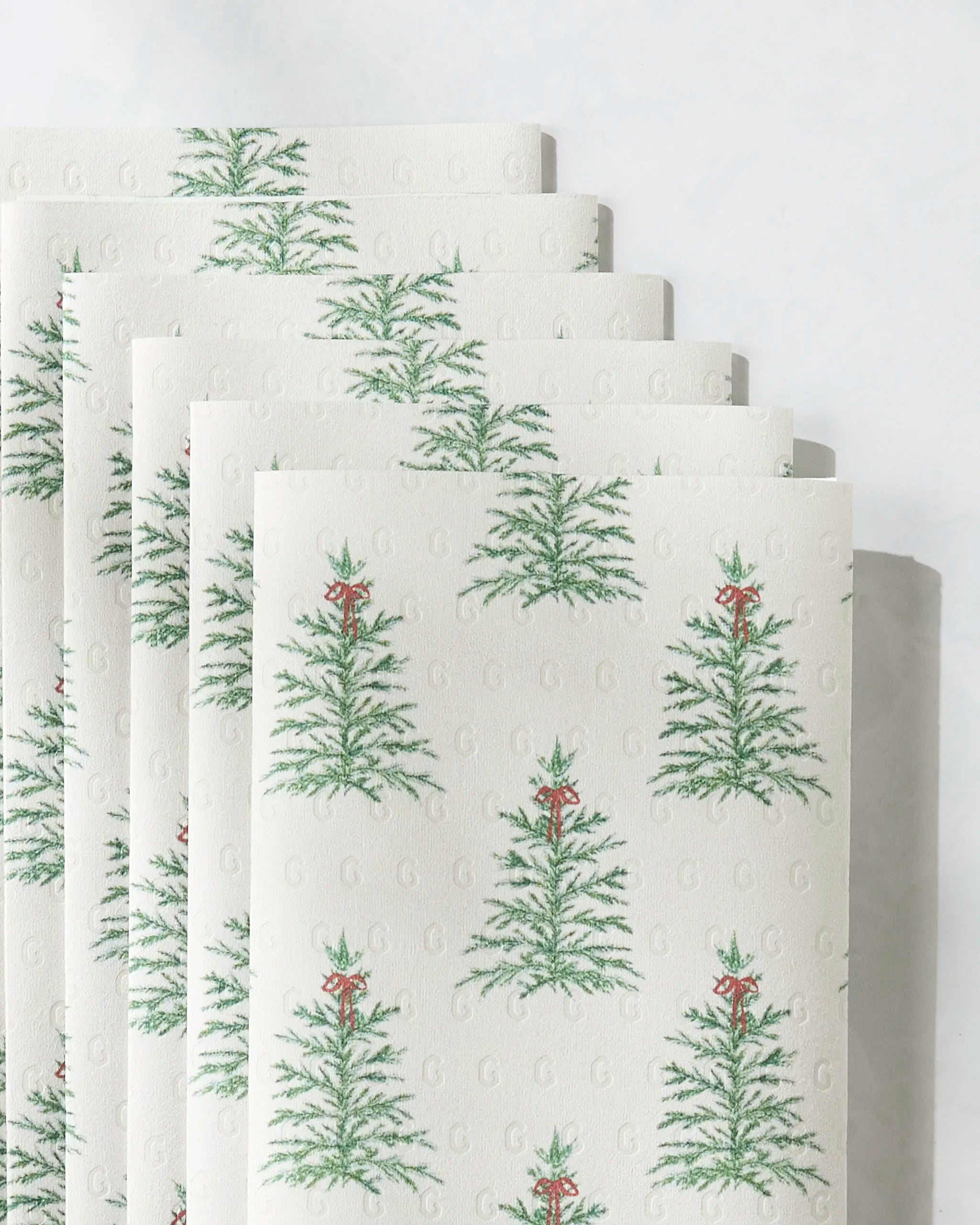 A close-up of Ellsworth eco-friendly paper towels by Geometry, featuring a repeating pattern of green Christmas trees with red decorations, stacked against a light, neutral background.