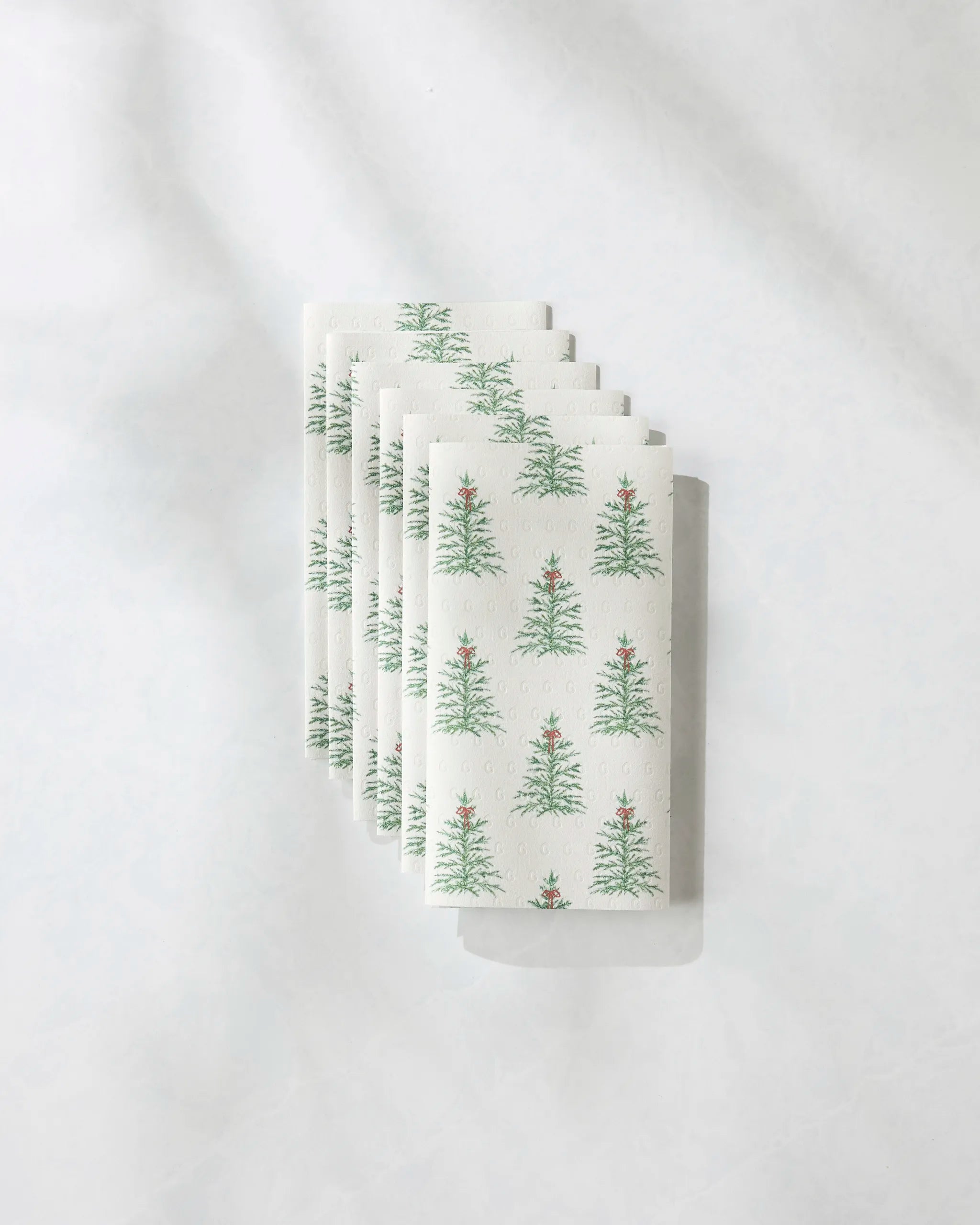 Five folded Geometry Ellsworth eco-friendly paper towels featuring a repeating Christmas tree pattern are stacked neatly on a white, textured surface.