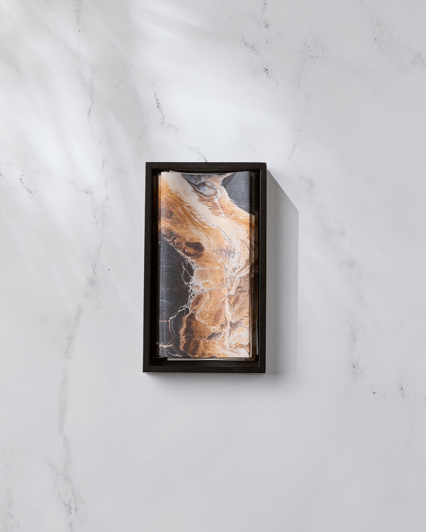 The Emperador by Geometry, featuring a rectangular black frame and an abstract brown, black, and white marble-inspired pattern, rests on a white marble surface with light gray veining as diagonal shadows fall across the scene.