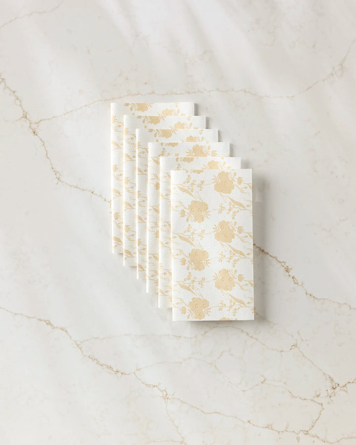 Eight white "Eternal" napkins by GEOMETRY, featuring a gold crab pattern and made from eco-friendly paper, are neatly stacked diagonally on a beige-veined white marble surface.
