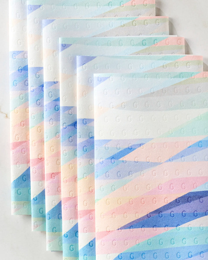 Five "Eventide" kitchen towels by Rachel Parker, featuring pastel diagonal stripes in blue, pink, and peach with embossed letter G patterns, are fanned out and slightly overlapping on a light surface.