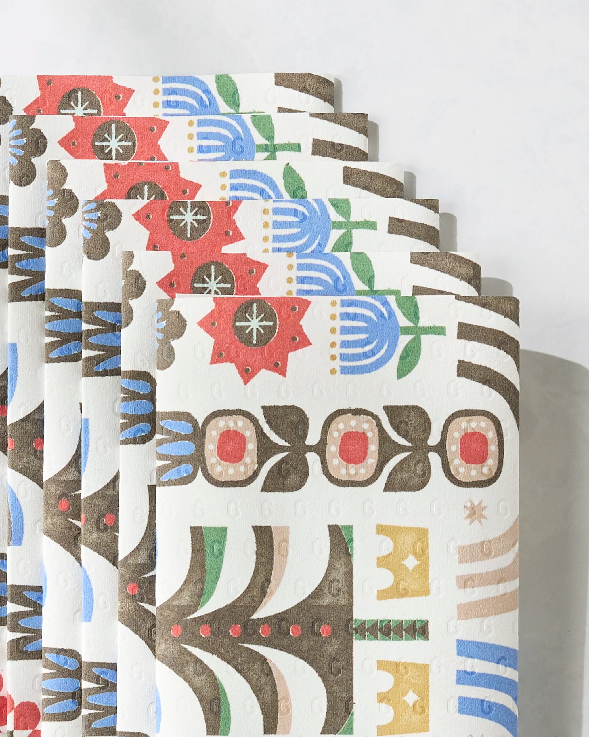 Everfolk kitchen towels by Geometry are eco-friendly, textured sheets stacked in a fan pattern, each adorned with vibrant red, blue, green, yellow, and brown folk art-inspired floral and geometric designs.