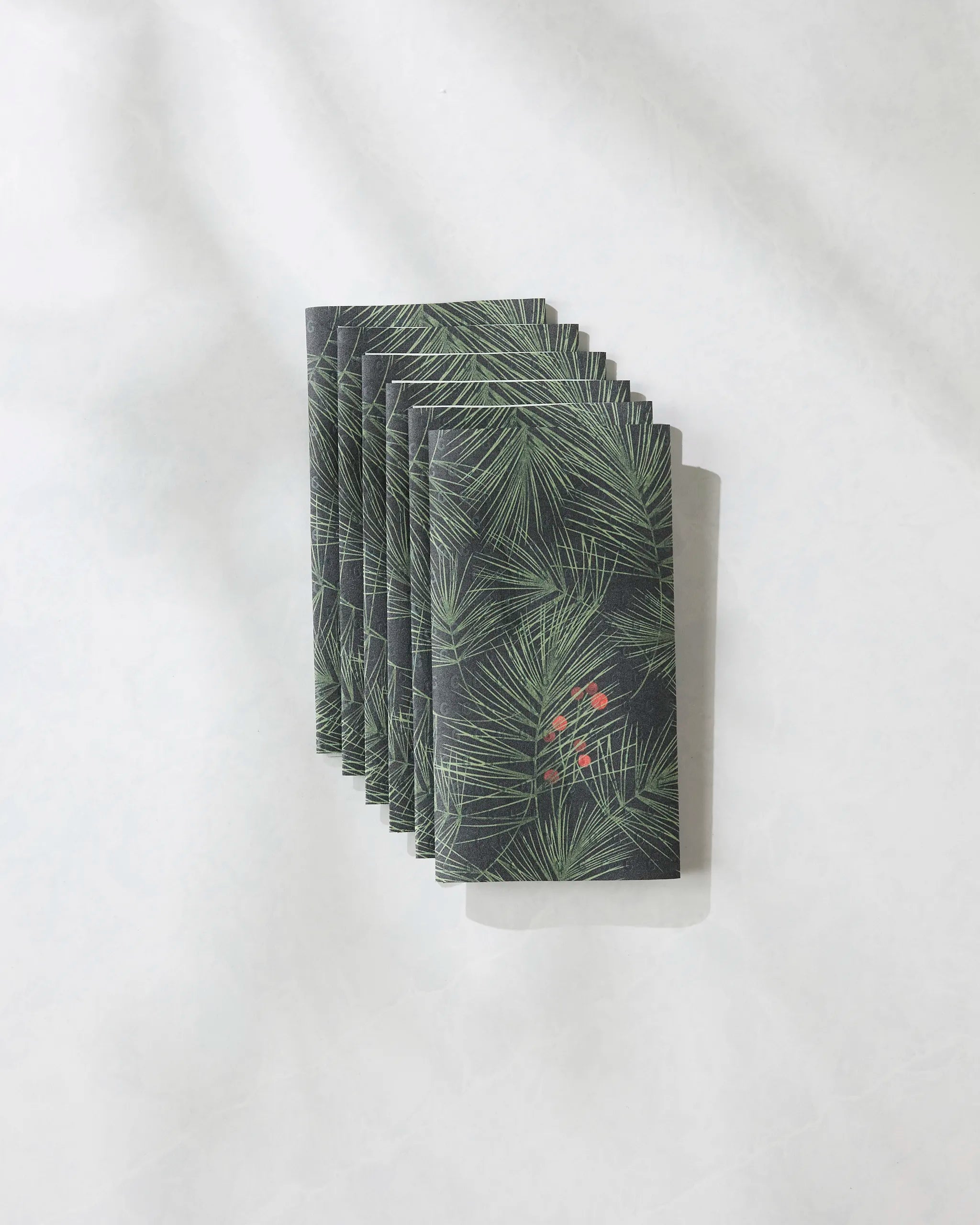 A stack of eight Geometry Evergreen Pine reusable paper towels, featuring a green pine needle and red berry pattern, is arranged neatly on a light, textured surface.