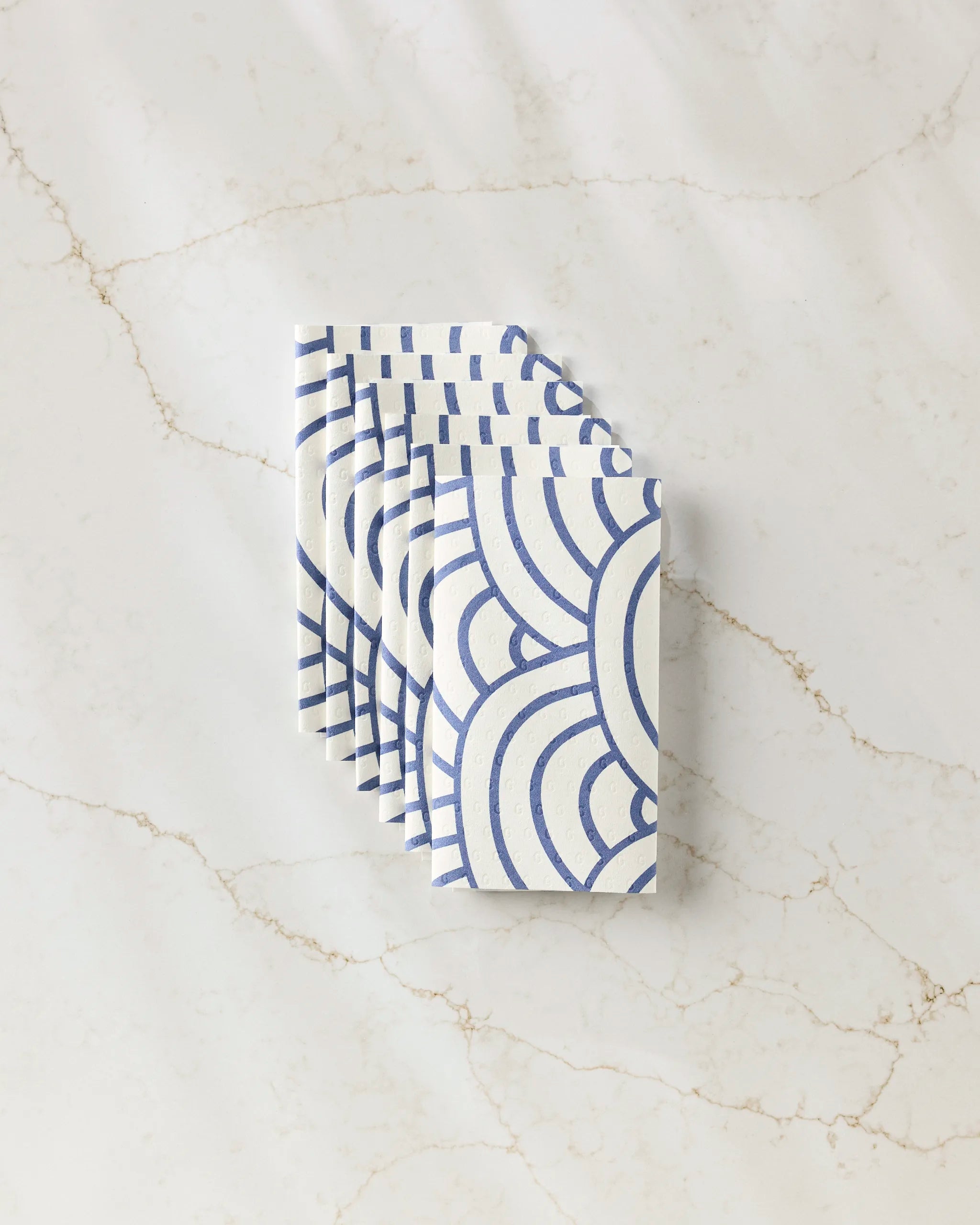 Six white Every Level reusable paper towels by GEOMETRY, featuring blue geometric curved line patterns, are neatly stacked on a light marble surface with beige veining—an eco-friendly choice for your table setting.