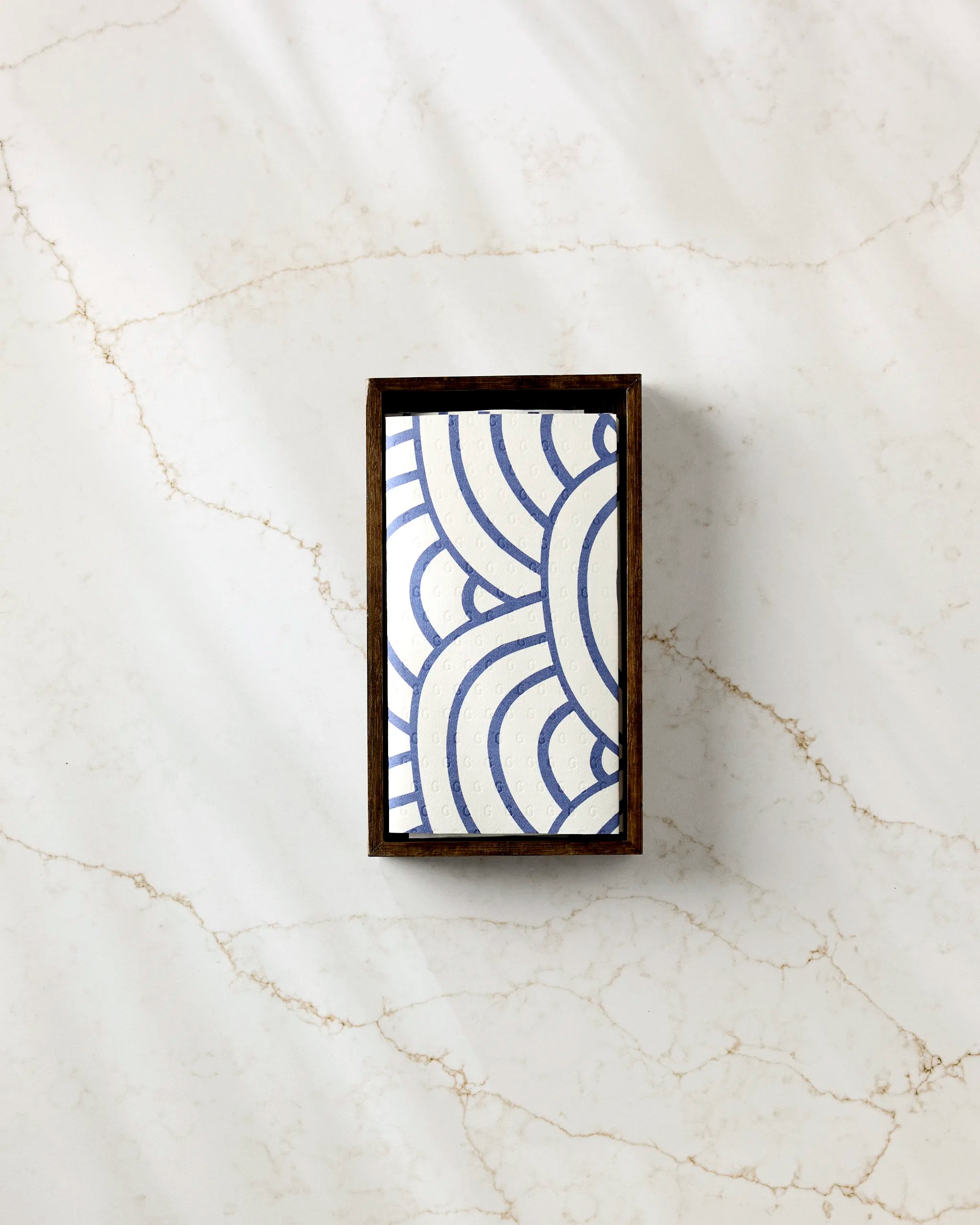 The Every Level bar soap by GEOMETRY, featuring a blue and white geometric wave design, rests in a dark wooden tray on light marble with brown veining, paired with eco-friendly paper towels for a sustainable accent.
