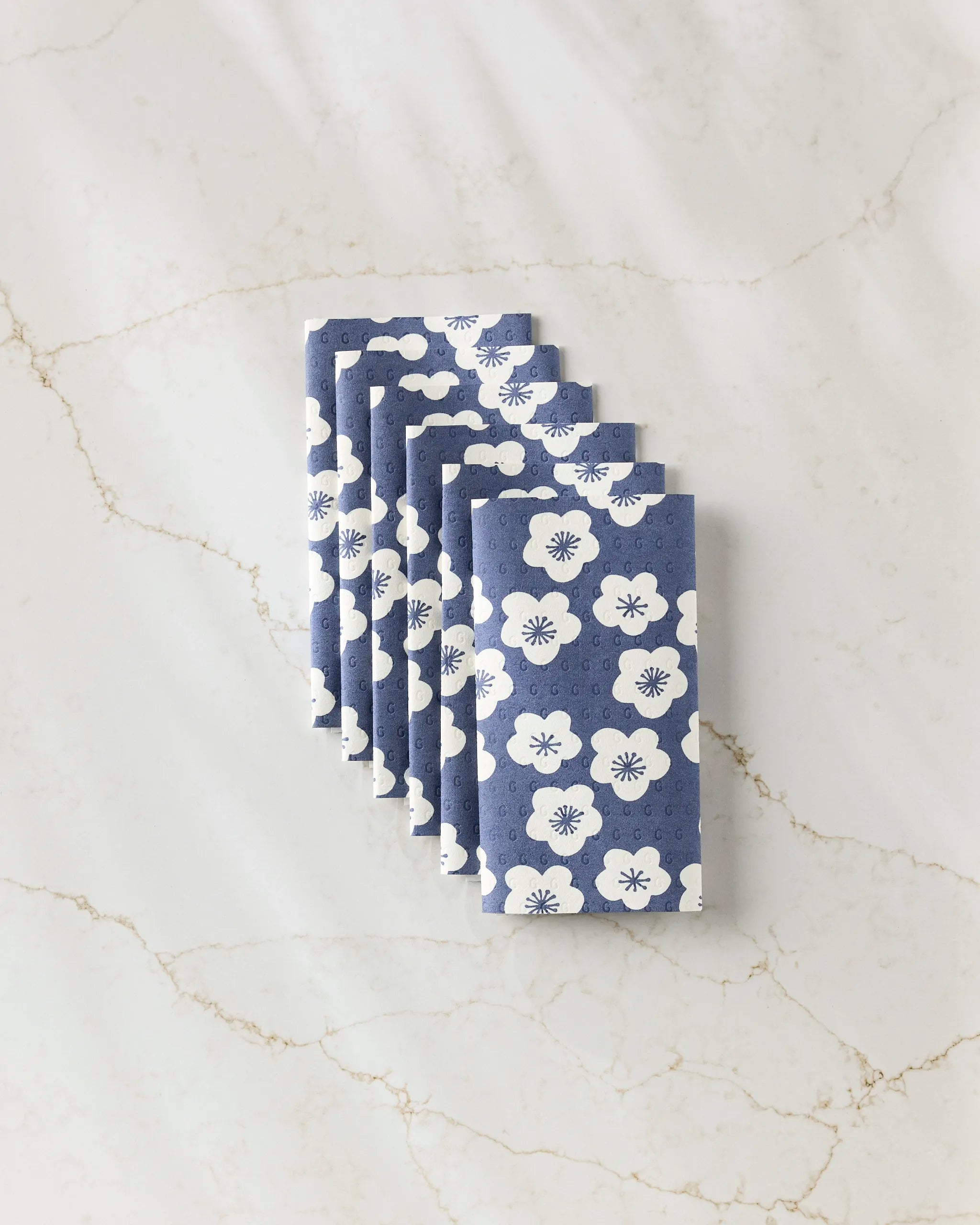 Six Every Sunday kitchen towels by GEOMETRY, blue with a white floral pattern and eco-friendly design, are neatly folded and arranged diagonally on a light marble surface.