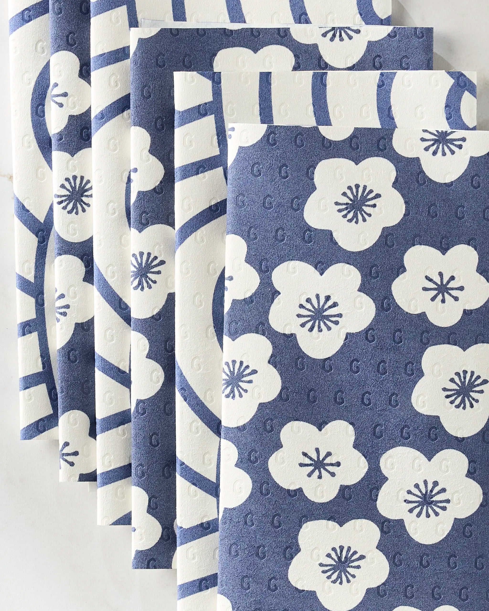 Six Every Wish kitchen towels by GEOMETRY are arranged in a fan shape, showcasing eco-friendly textured fabric with blue and white five-petal floral and abstract patterns on alternating blue and white backgrounds.