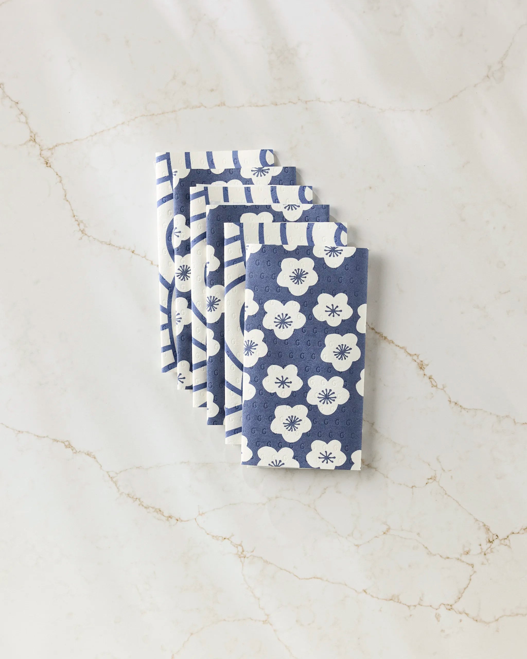 Four folded blue and white GEOMETRY Every Wish eco-friendly kitchen towels with floral and striped designs are neatly arranged on a white marble surface with light brown veining.