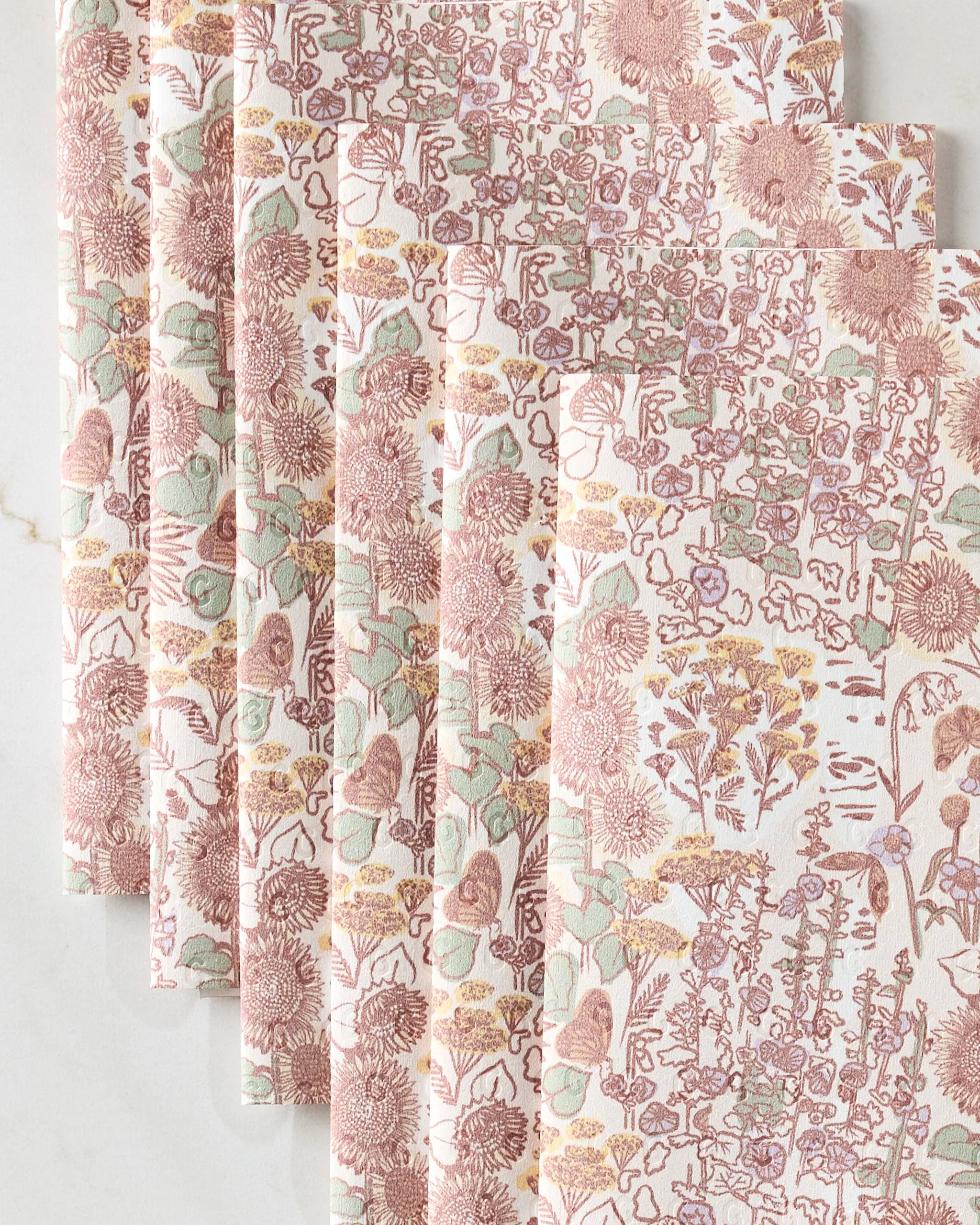 Five folded, reusable paper towels from Elisabeth Wing’s Fall Forest collection are stacked on a light marble surface. Their intricate floral and leafy design in pink, yellow, and green adds eco-friendly style to your kitchen.