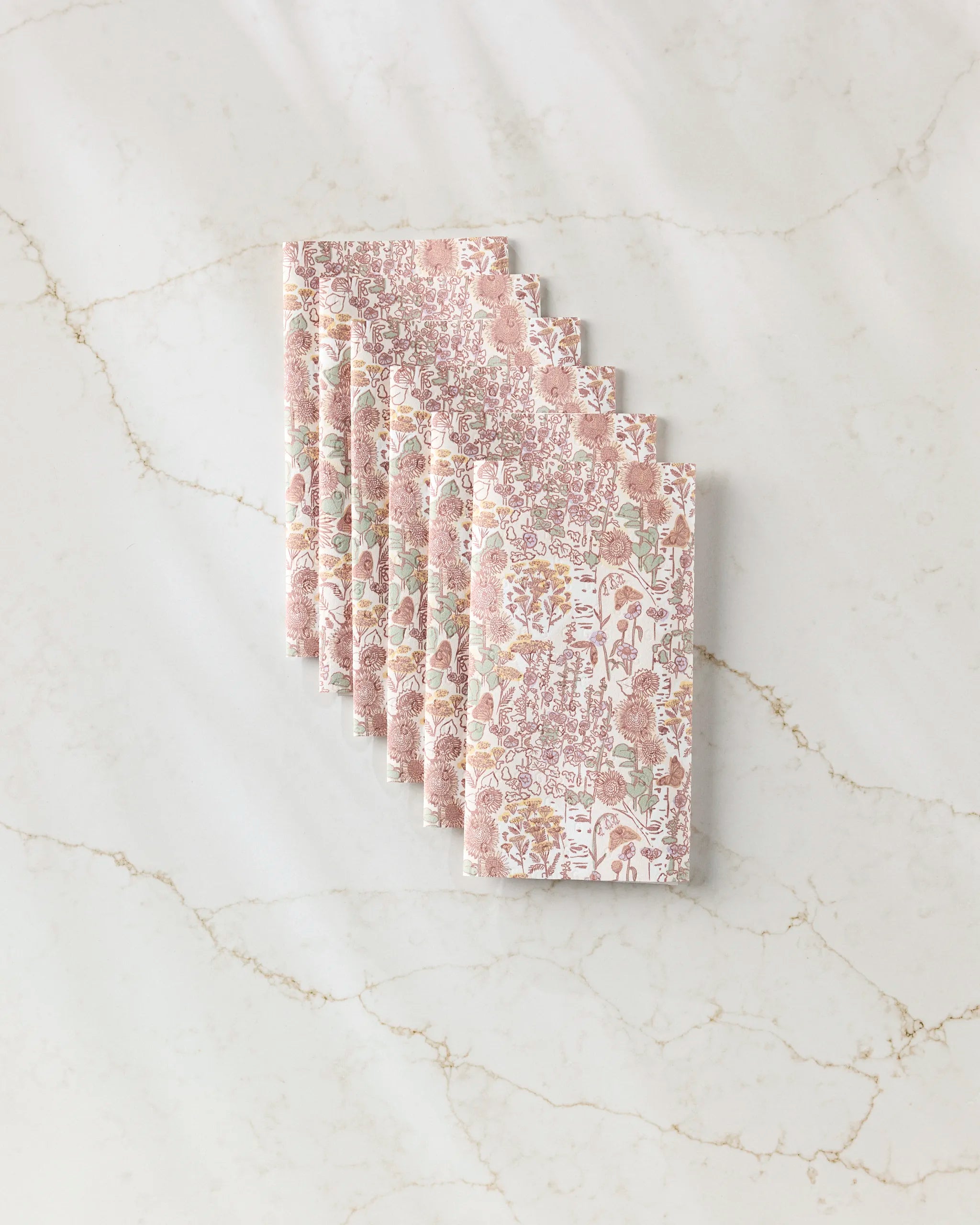 Six folded Fall Forest napkins by Elisabeth Wing, featuring pink, green, and beige florals, are fanned on white marble—ideal as eco-friendly kitchen towels or reusable paper towels for sustainable living.