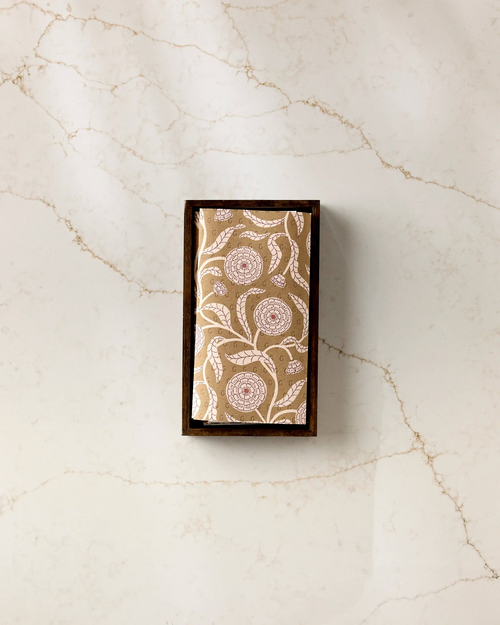 The Falling Flowers soap by Teri the Butter, a rectangular bar with a floral pattern, rests in a dark wooden tray on light marble with beige veining next to a stack of eco-friendly kitchen towels.