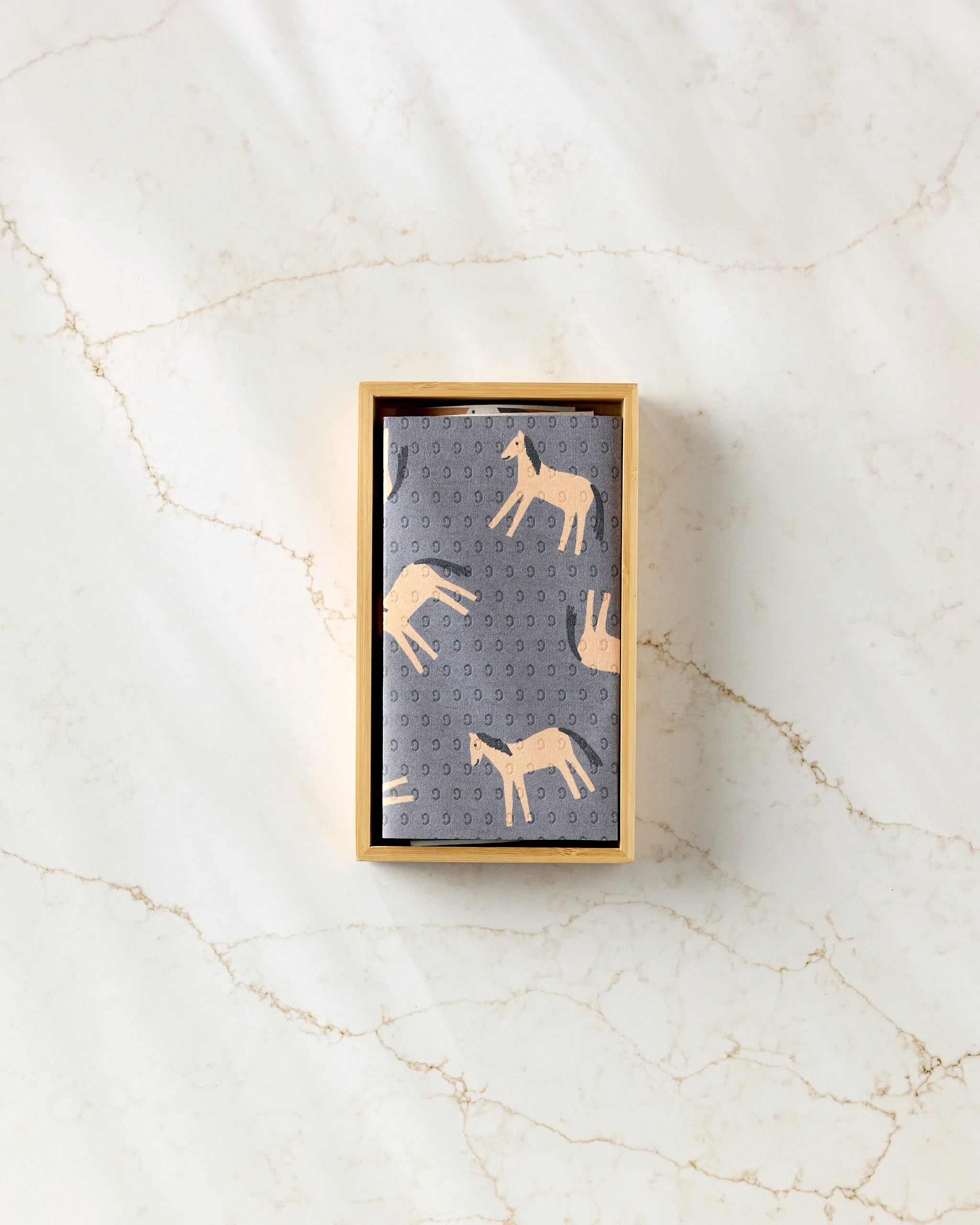 A rectangular wooden box holds a folded grey "Farm Animals" cloth by Coit Creative—an eco-friendly paper towel alternative featuring beige animal illustrations—set on white marble with light brown veining.