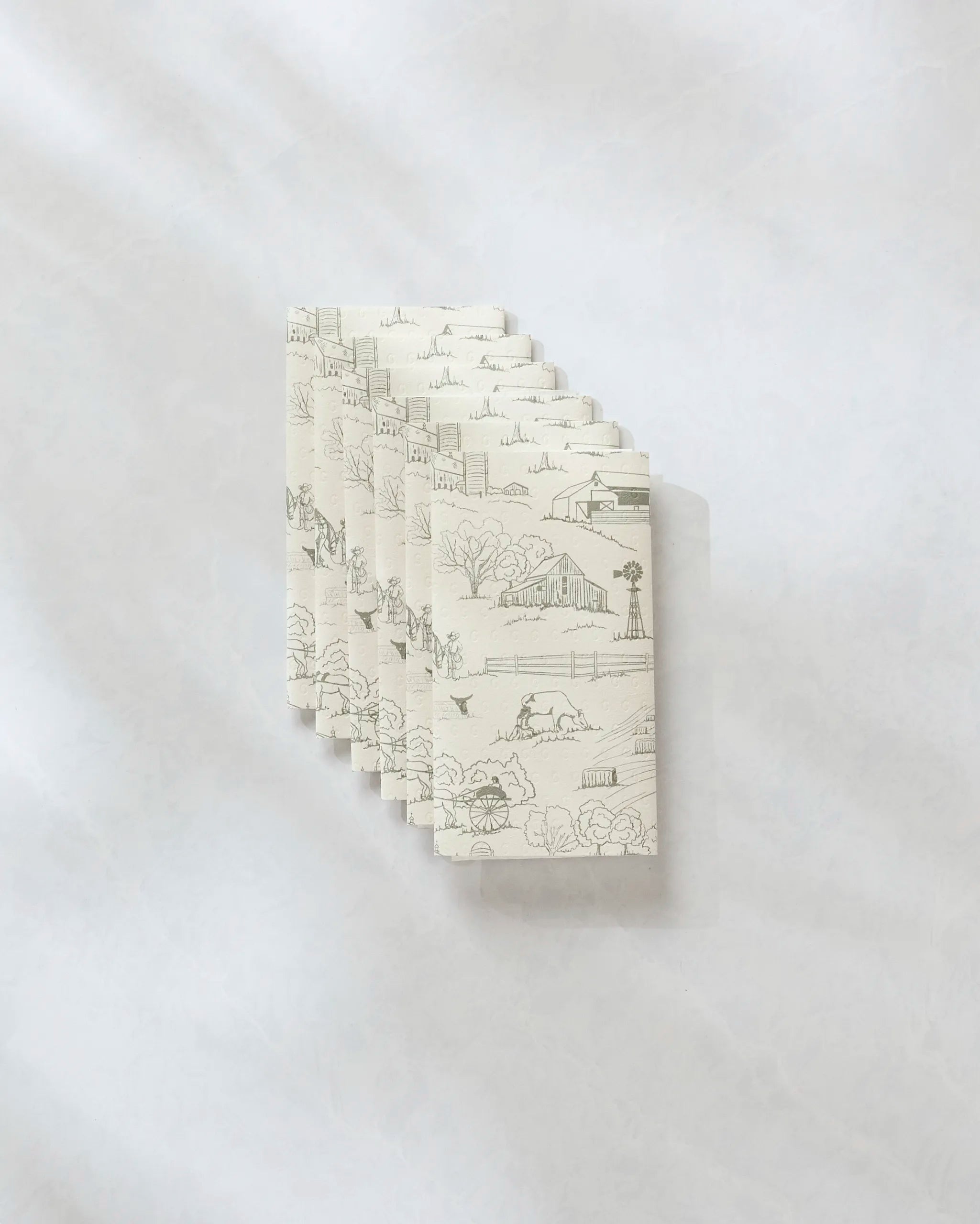 A stack of folded white Farmyard reusable paper towels by Hannah Langford, decorated with gray barns, trees, and windmills, arranged on a pale marbled surface.