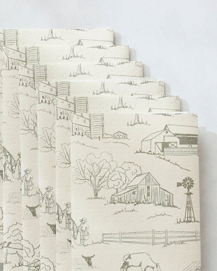 Several sheets of Hannah Langford’s Farmyard eco-friendly, off-white paper towels, featuring printed rural scenes with barns, trees, fences, a windmill, and people with animals, are layered neatly against a light background.