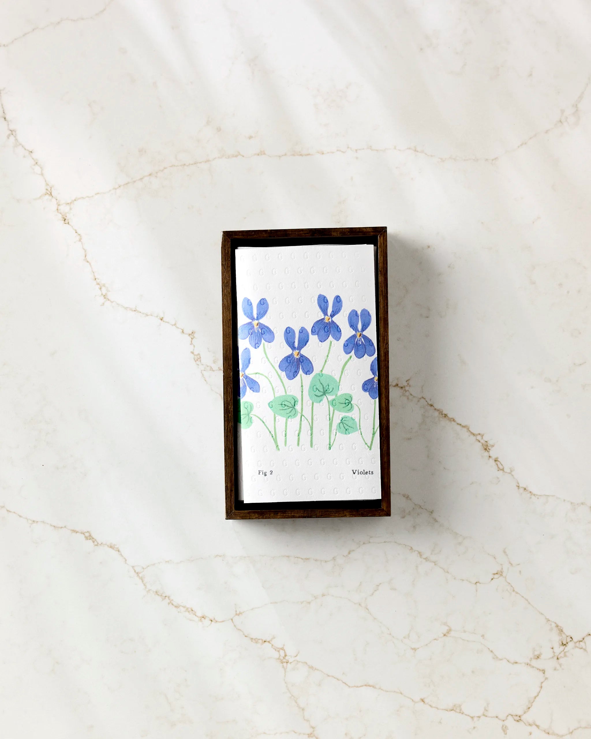 The February Violets by Linda Fox is a small box with a white lid, hand-painted with blue and green flowers, perfect for storing eco-friendly paper towels. It sits beautifully on any marble surface with subtle gray and tan veins.