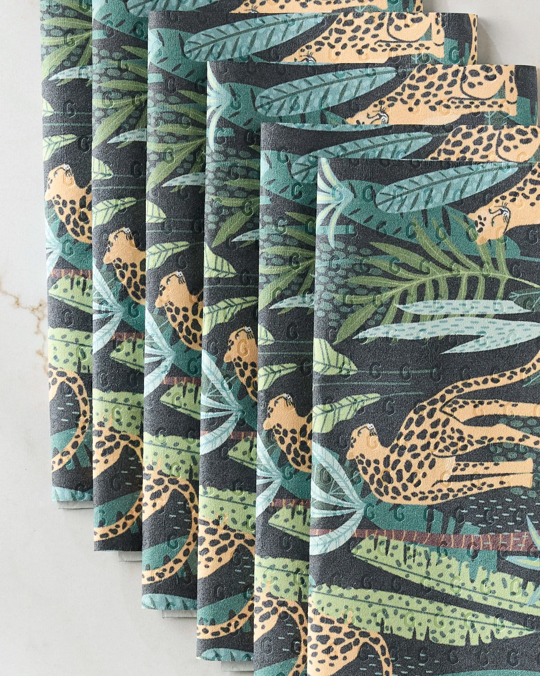 GEOMETRY's Feeling Spotty cloth napkins, showcasing a lively jungle design with leopards and green leaves, are arranged on white marble—a stylish, eco-friendly alternative to paper towels.