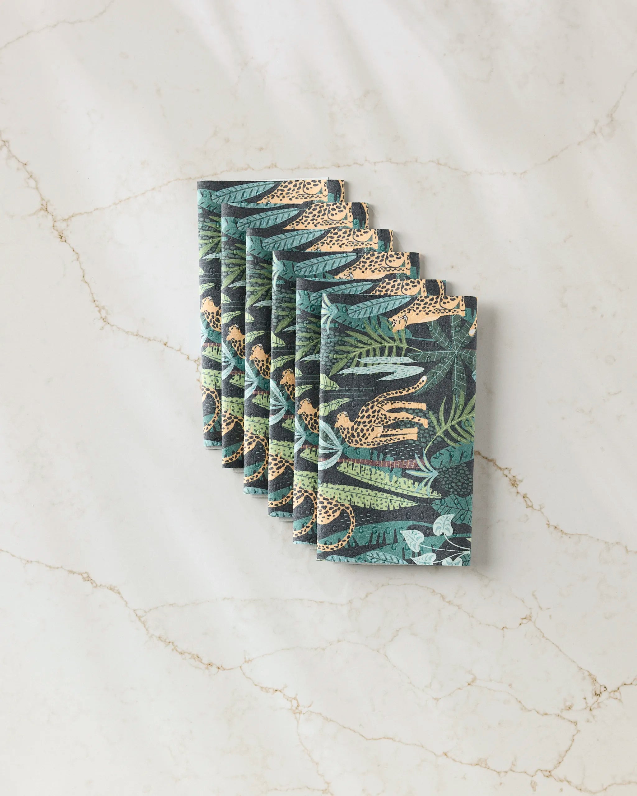 A neatly arranged stack of GEOMETRY Feeling Spotty eco-friendly paper towels, showcasing vibrant leopards, green leaves, and jungle foliage, sits on a white marble surface with light brown veins.