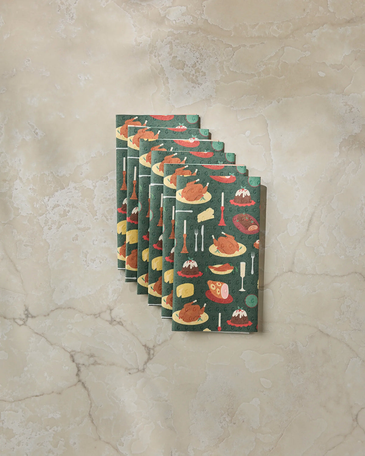 Five Festive Feast napkins by Geometry, crafted from eco-friendly paper towels and decorated with a Thanksgiving-themed print of turkeys, ham, and side dishes, are folded and fanned out on a beige marble surface.