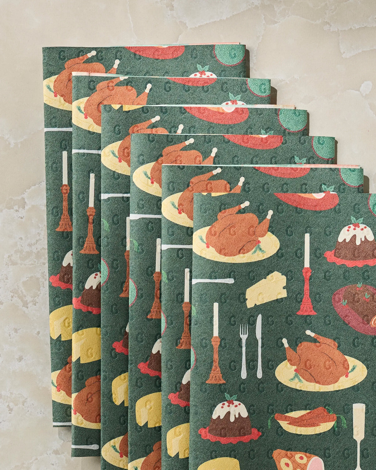 Five Geometry “Festive Feast” eco-friendly paper towels, featuring a green background adorned with illustrations of roast turkey, cheese, pudding, plates, cutlery, and candlesticks, are arranged in a fan shape on a marble surface.