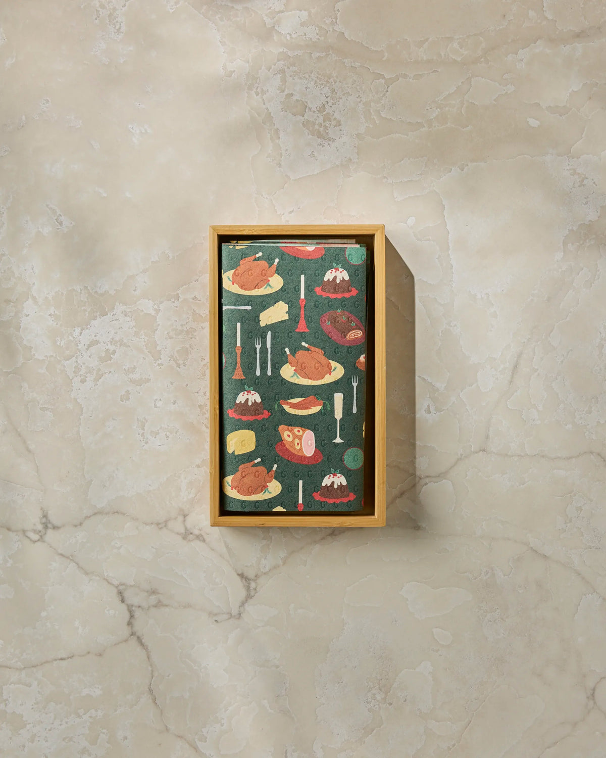 The “Festive Feast” rectangular wooden tray by Geometry, adorned with a vibrant food-themed design featuring illustrations of meats, bones, and side dishes, rests elegantly on a light marble surface and is paired perfectly with eco-friendly paper towels for sustainable dining.