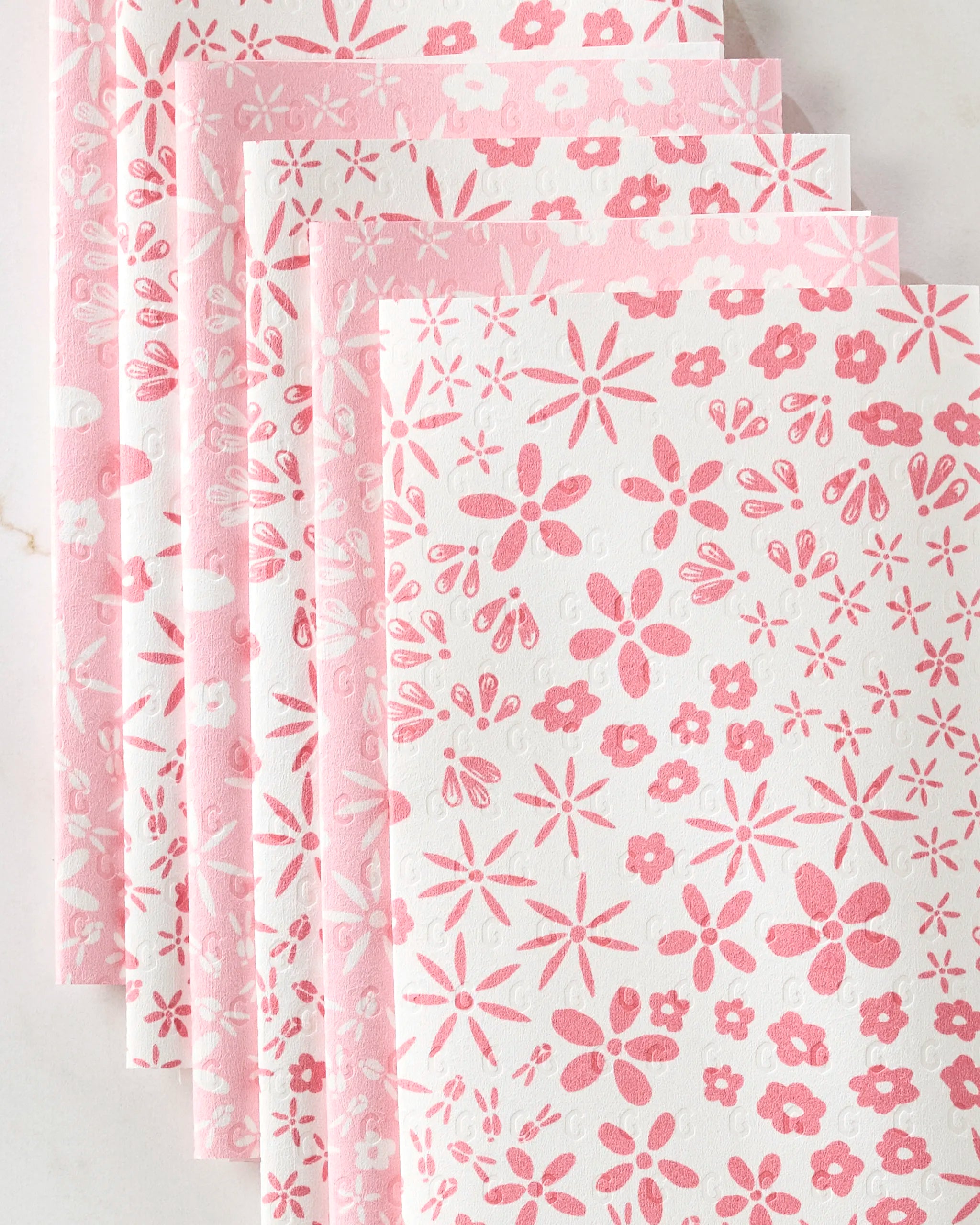 Six eco-friendly Fields of Flowers kitchen towels by Hufton Studio are fanned out, each showing pink flower and starburst patterns on a white background.