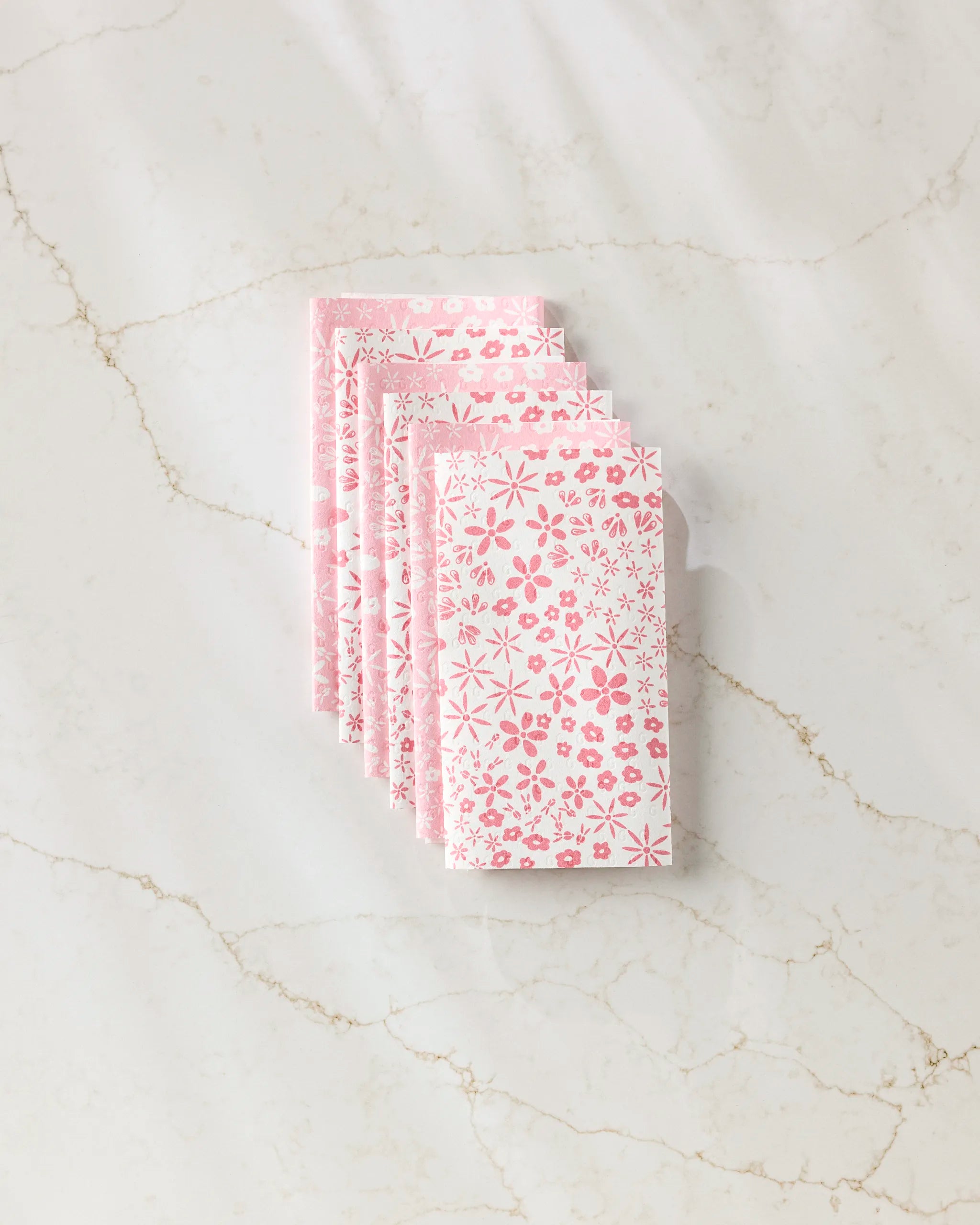 Four folded Fields of Flowers cloth napkins by Hufton Studio, with a pink and white floral pattern, are neatly stacked on marble—offering a charming, eco-friendly alternative to disposable paper towels.