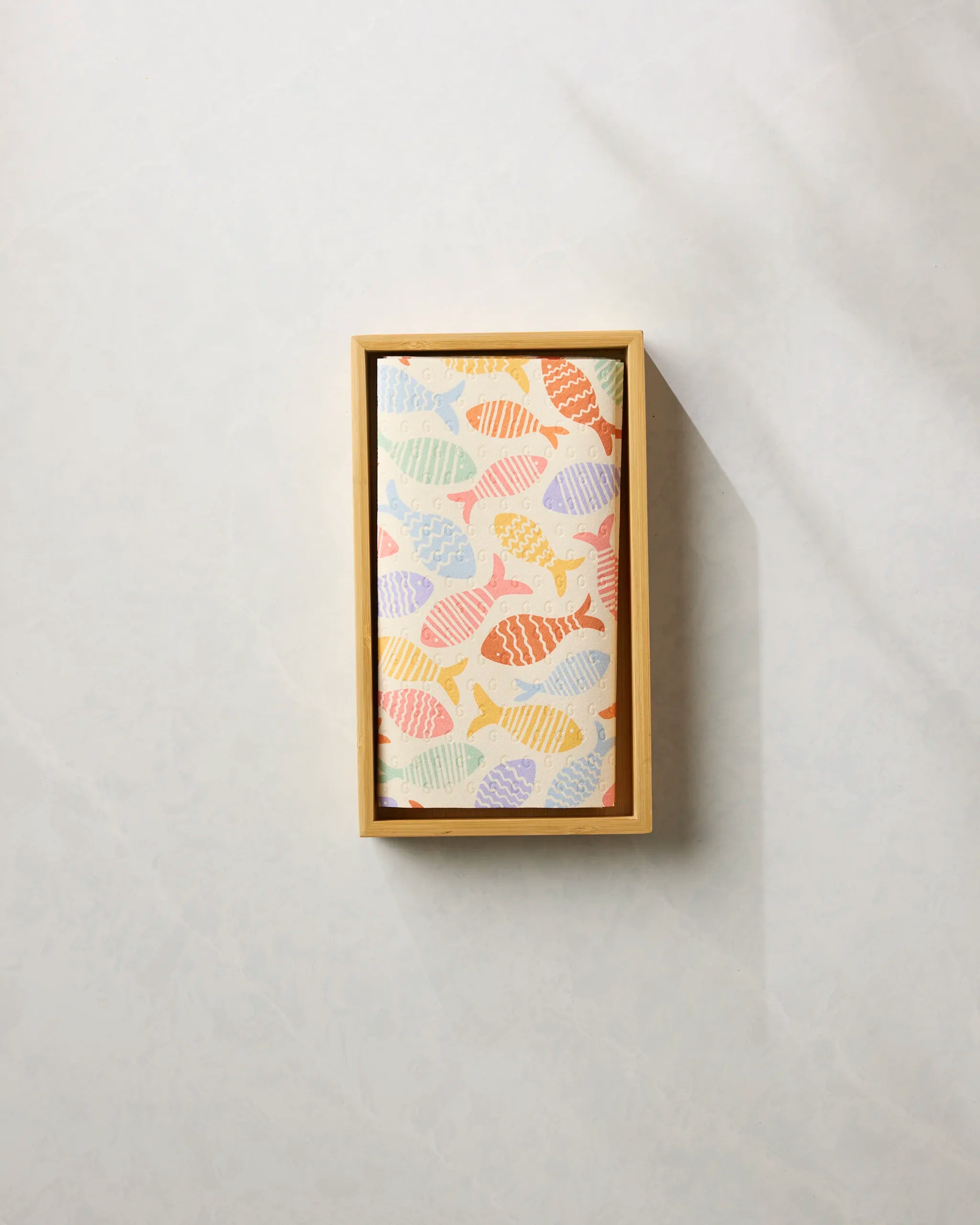 The Geometry Fish Flip is an eco-friendly kitchen towel and recycled paper towel alternative, featuring a colorful fish pattern and presented in a rectangular wooden box on a marbled surface.