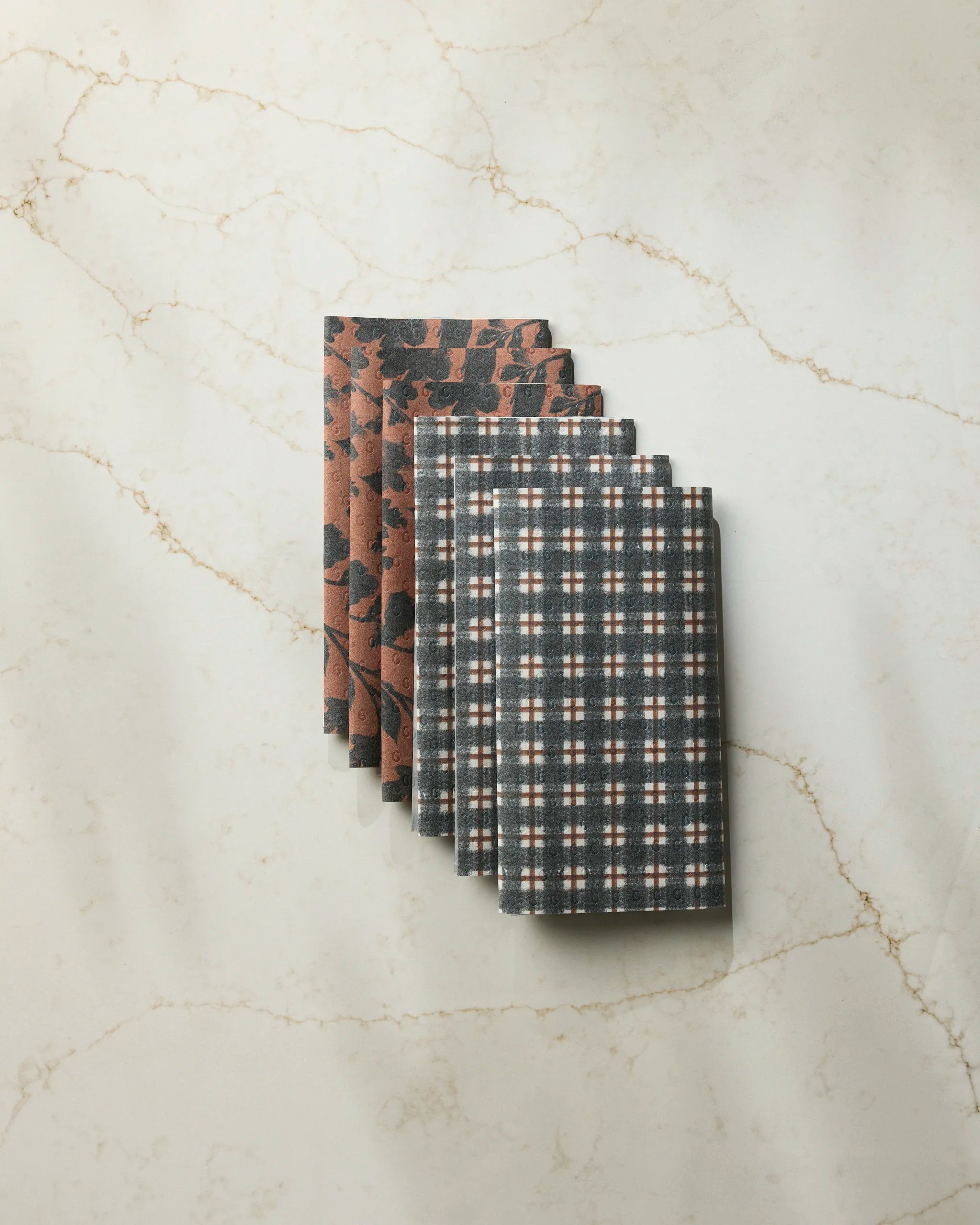 Two folded Flint Hallowell kitchen towels by Geometry rest on a light marble surface. The top towel displays a black and rust geometric pattern; the bottom shows a black, white, and rust plaid design.