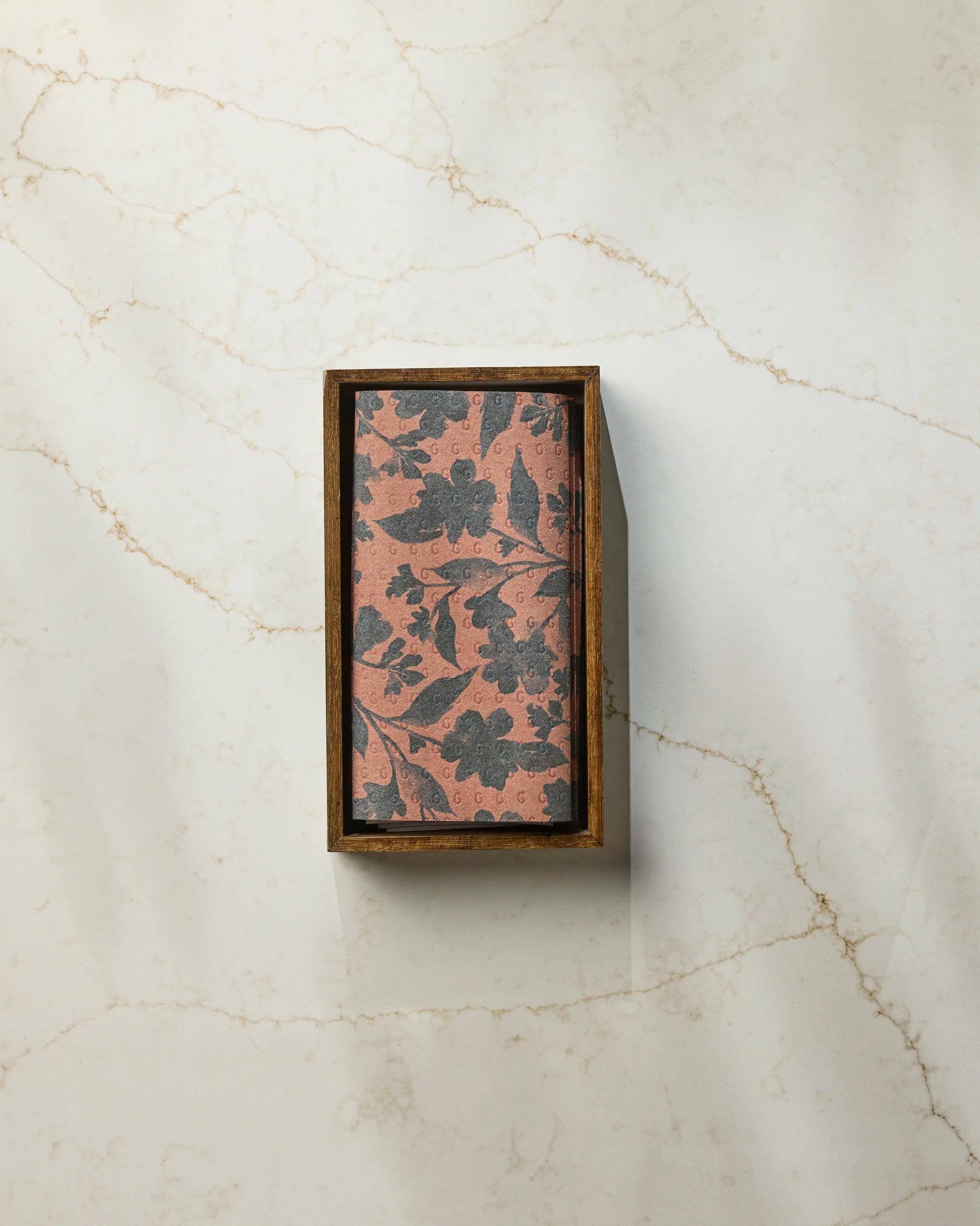 The Flint Hallowell soap by Geometry, featuring a pink base and dark floral pattern, sits in a wooden box on white marble with light brown veins, next to folded reusable paper towels for an eco-friendly touch.