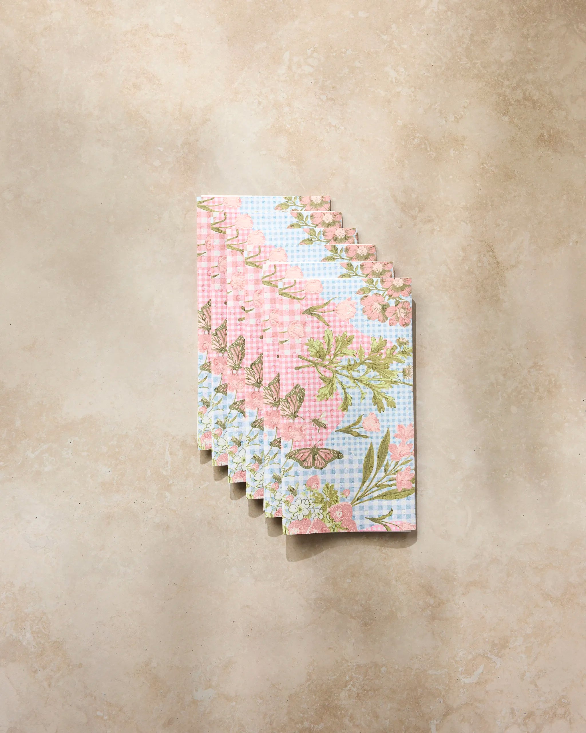 Six eco-friendly Geometry “Floral Collage” paper towels, featuring pink, green, and white floral and checkered patterns, are fanned out on a light beige marble surface.