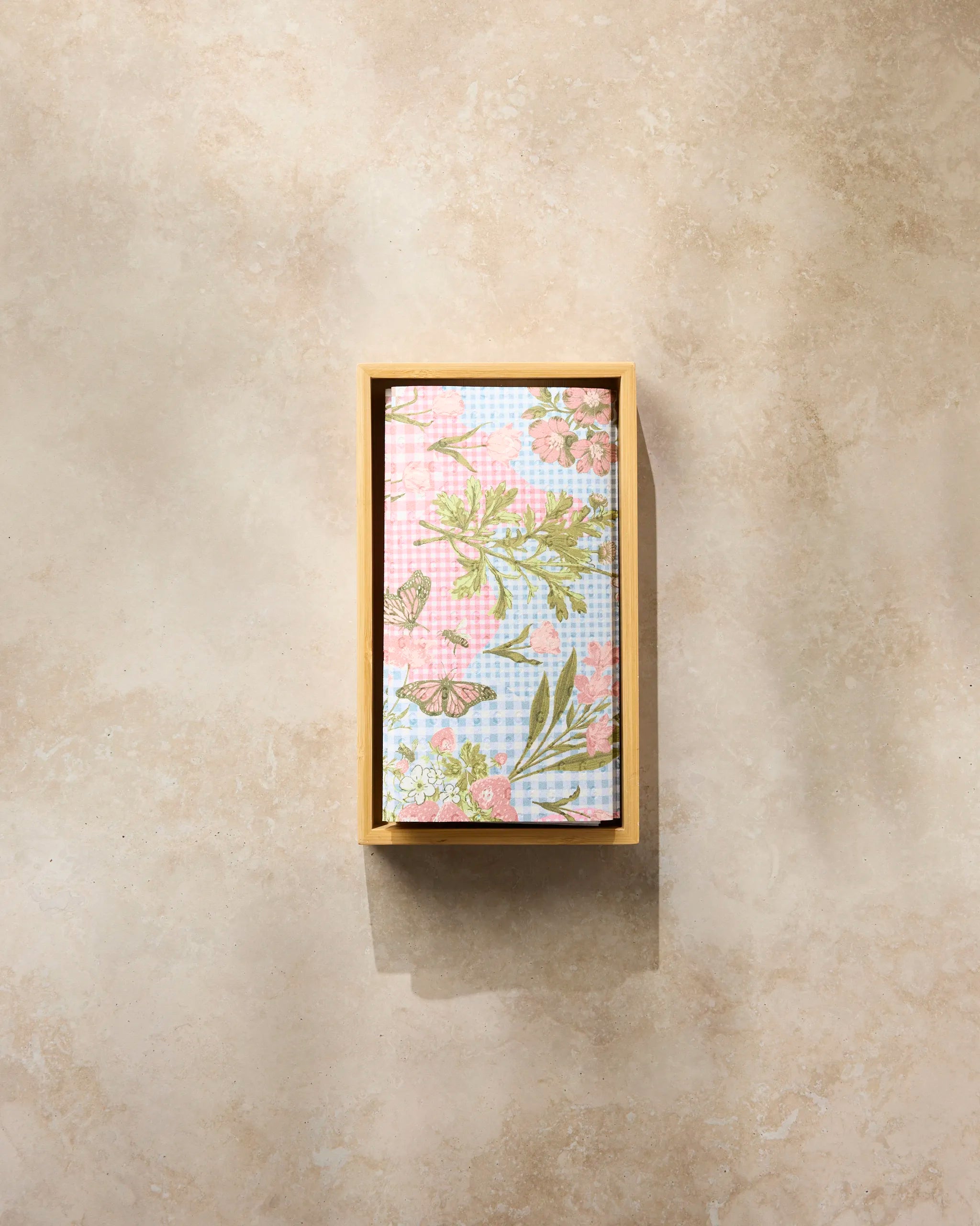 The Geometry Floral Collage reusable paper towel, featuring a pink, green, and light blue floral pattern, is folded neatly inside a rectangular wooden box on a beige textured surface.