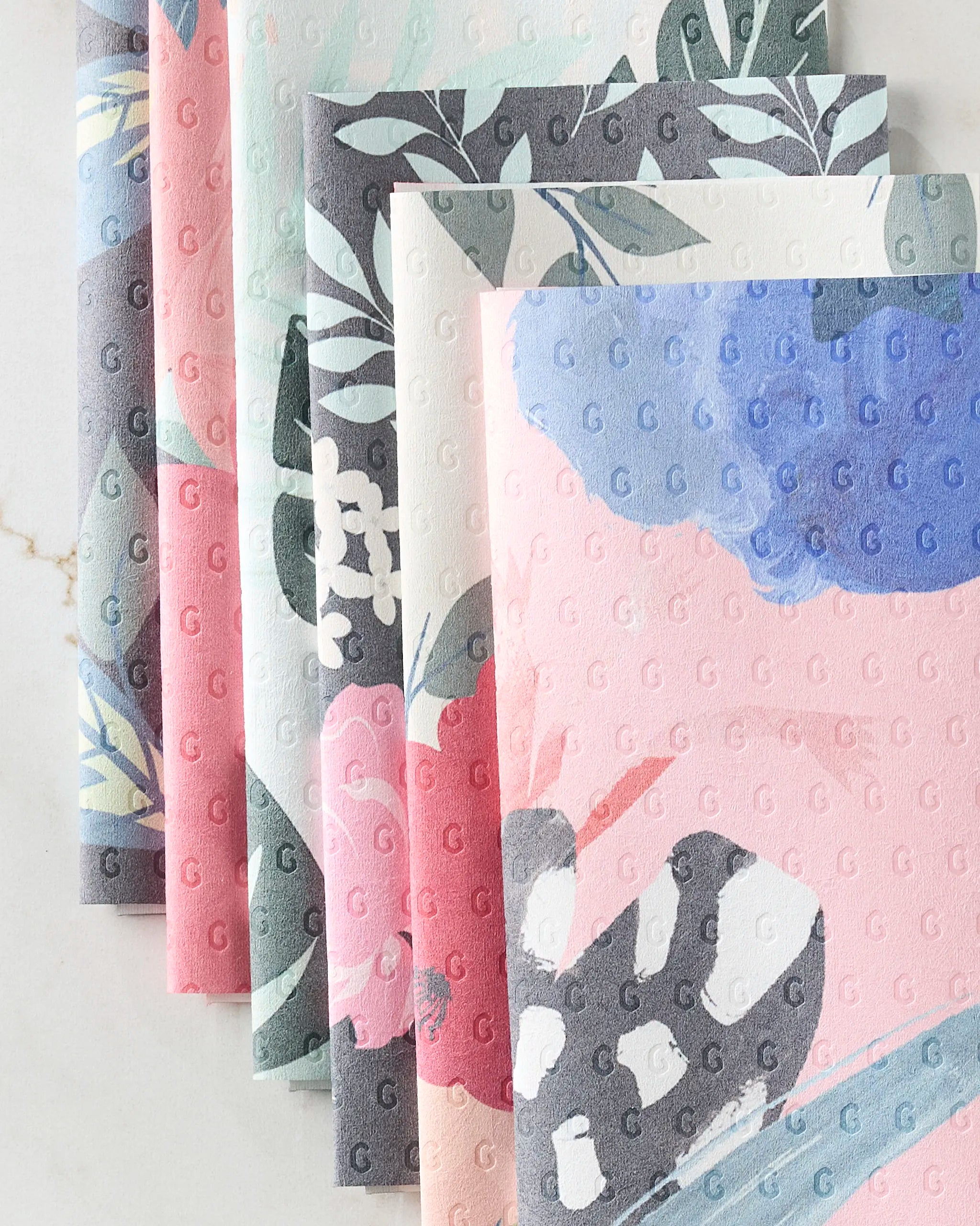 Five folded Floral Fun reusable towels by GEOMETRY, featuring pastel floral and leaf prints in pink, blue, gray, and white, are fanned out on a light surface. These stylish towels offer a sustainable alternative to disposable paper products.