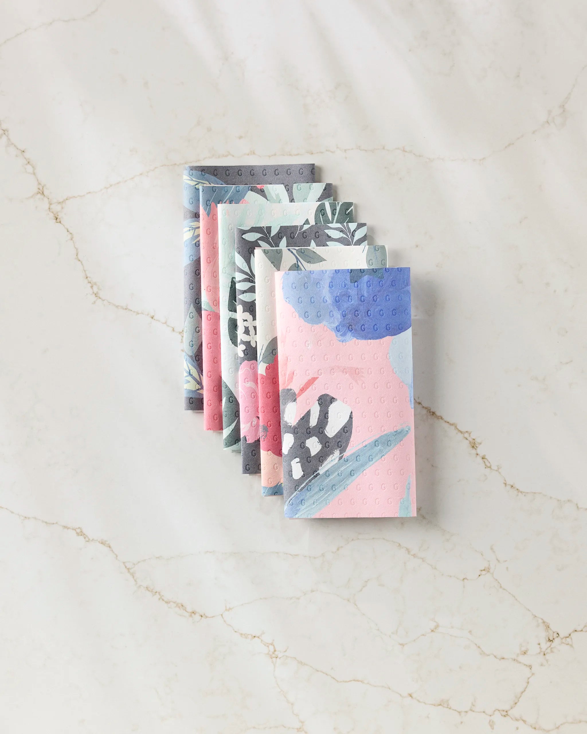 Six GEOMETRY Floral Fun reusable paper towels with abstract floral patterns in pastel pink, blue, and gray are neatly arranged in a row on a white marble surface.