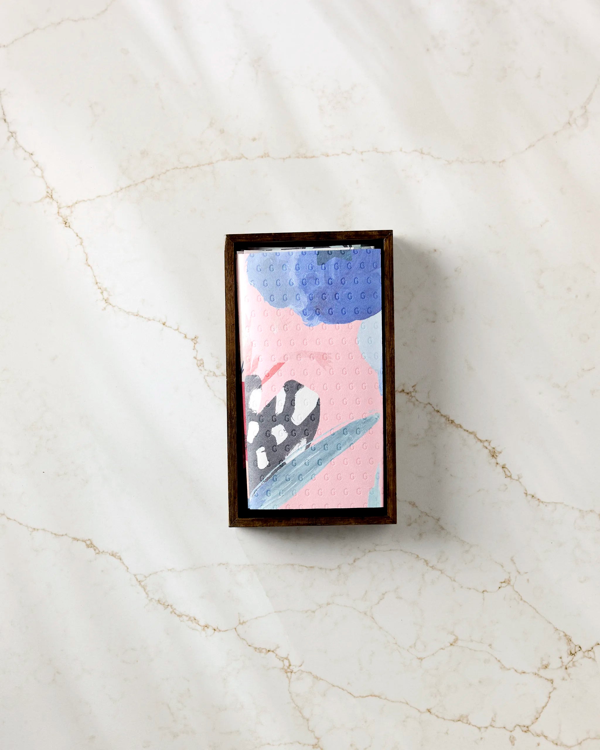 The GEOMETRY Floral Fun bar soap, featuring a pink, blue, and gray abstract design, sits in a brown wooden dish on white marble next to a roll of eco-friendly paper towels.