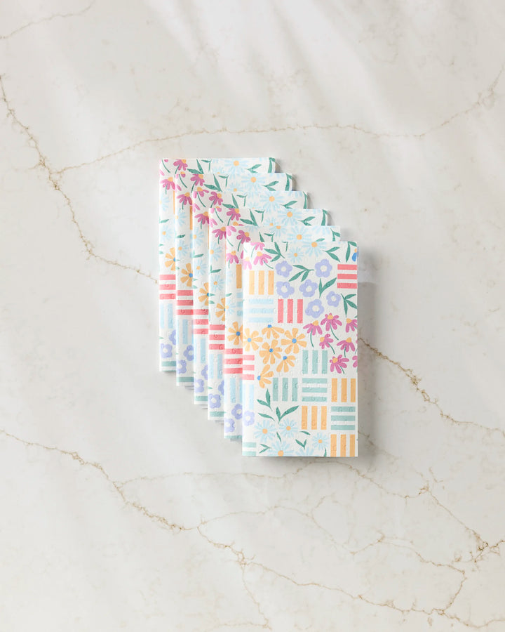 Six "Floral Garden" cloth napkins by Charlie Rowan Designs, featuring vibrant floral and geometric patterns inspired by reusable paper towels, are neatly fanned out on a white marble surface.