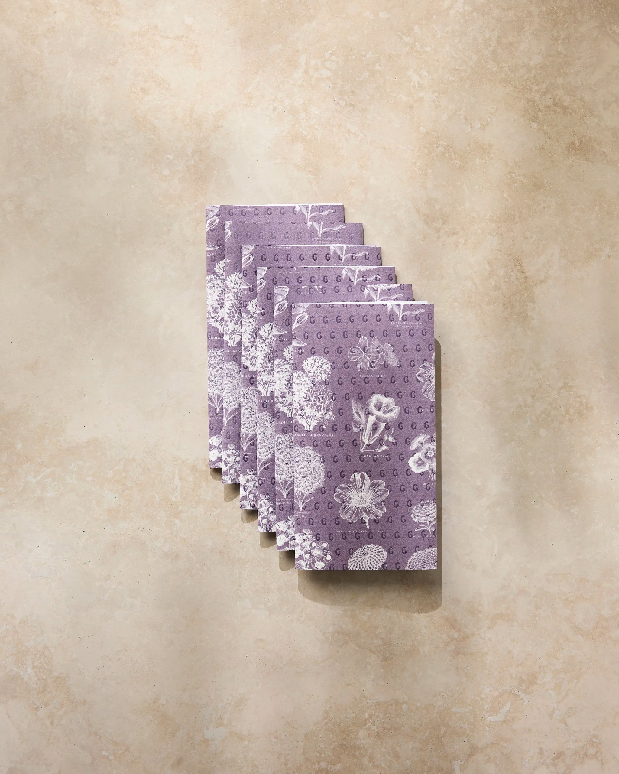 Six Geometry “Flower Glossary White” envelopes with white botanical designs are fanned out on beige marble—a stylish choice for gifting eco-friendly kitchen towels or other sustainable treasures.