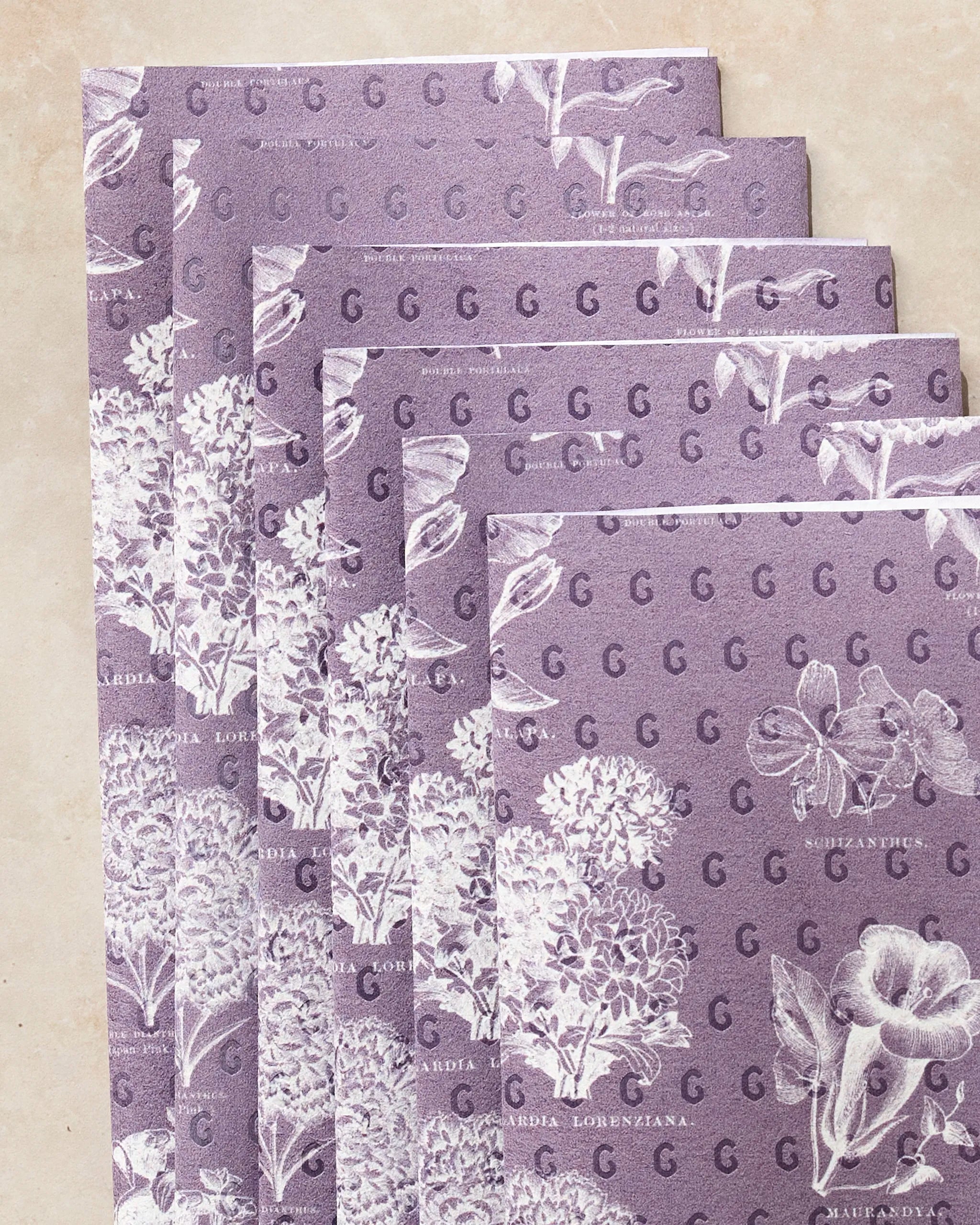 Five stacked sheets of Geometry’s Flower Glossary White eco-friendly towels feature a repeating pattern of white botanical illustrations and the letter G. Each sheet has decorative flowers and leaves in unique arrangements.