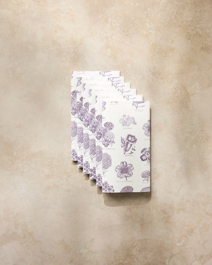 Seven compact, white "Flower Glossary" booklets by Geometry, featuring purple floral designs, are neatly stacked on a light beige textured surface.