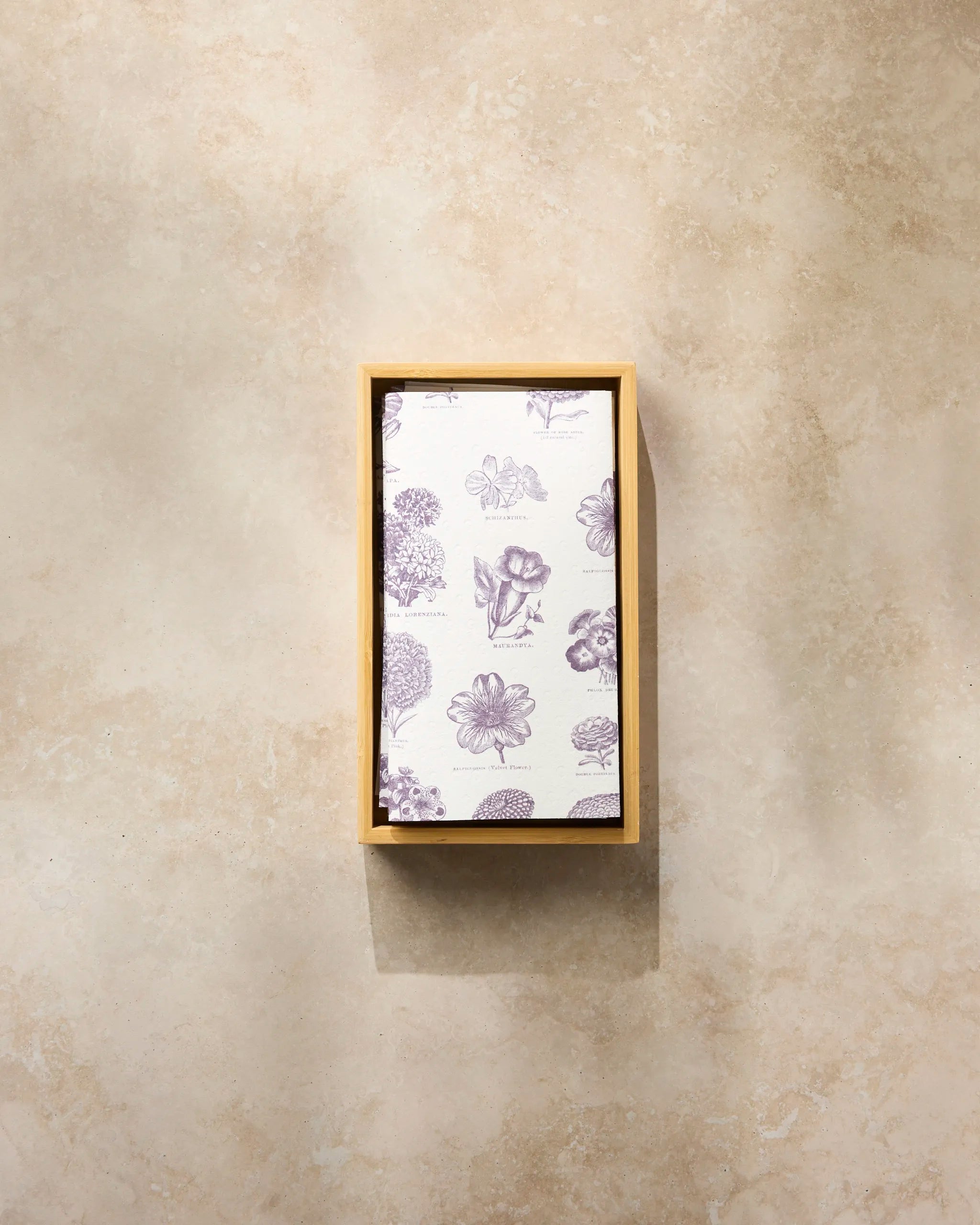 A rectangular wooden tray holds Geometry's Flower Glossary eco-friendly kitchen towel, featuring a white cloth with a purple floral pattern, placed on a light beige textured surface.