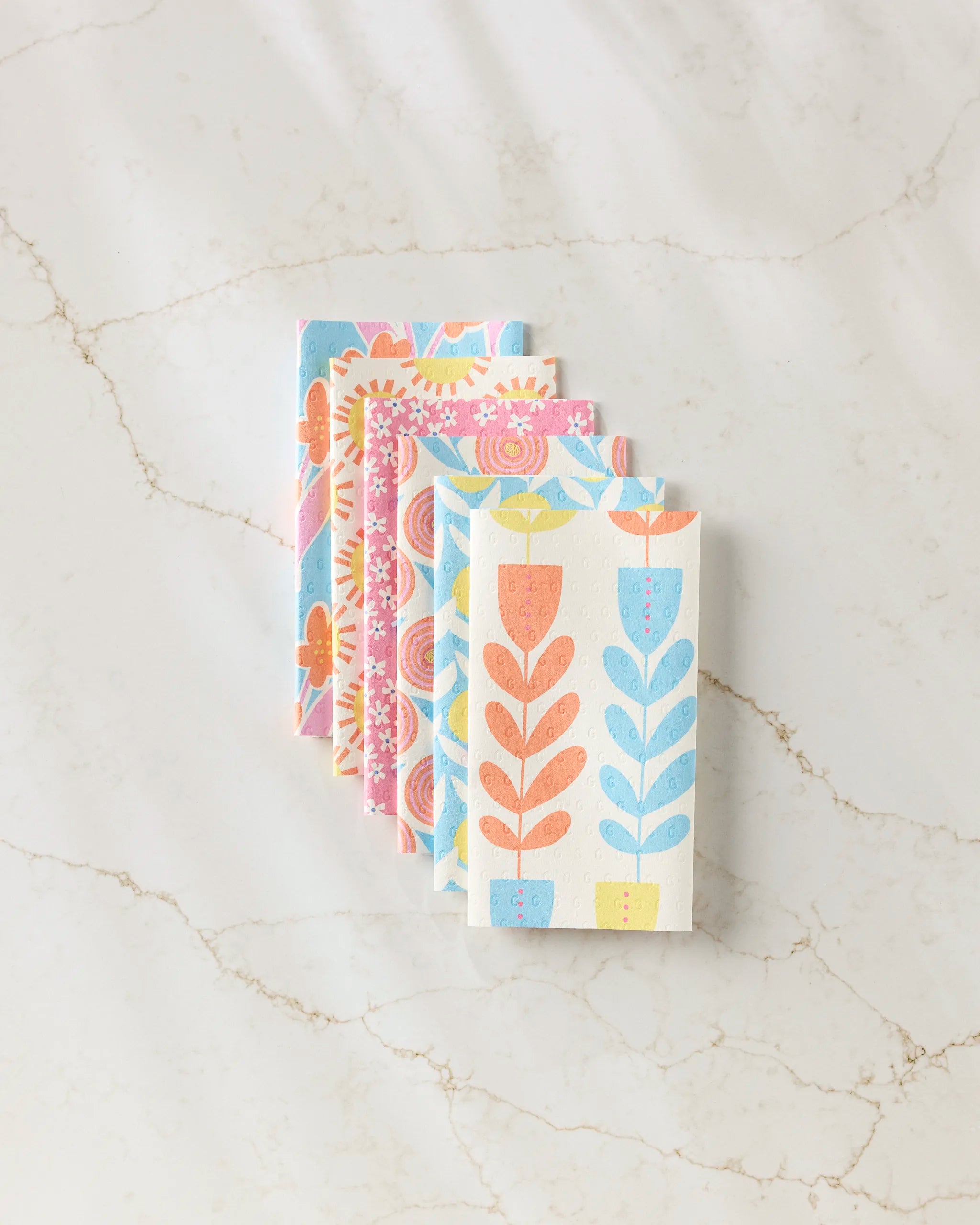 Five reusable Flower Parade paper towels by Lizzie Clark, featuring pastel floral and leaf designs in blue, pink, and orange, are stacked on a white marble surface with light brown veining.