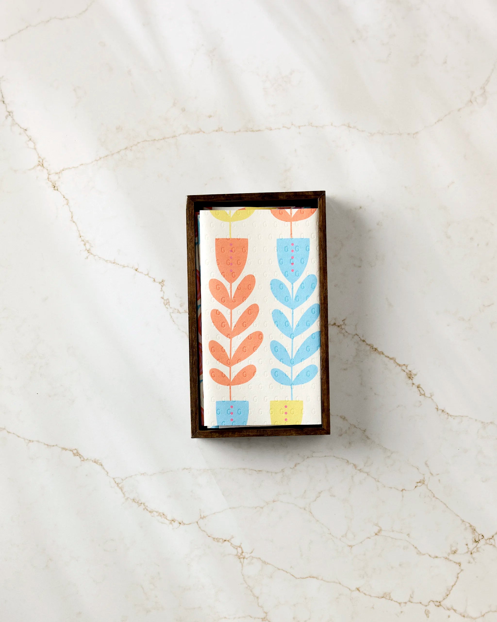 The Flower Parade soap bar by Lizzie Clark, featuring stylized orange, yellow, and blue flowers, rests in a dark wooden dish on white marble with tan veins, next to a stack of eco-friendly paper towels.