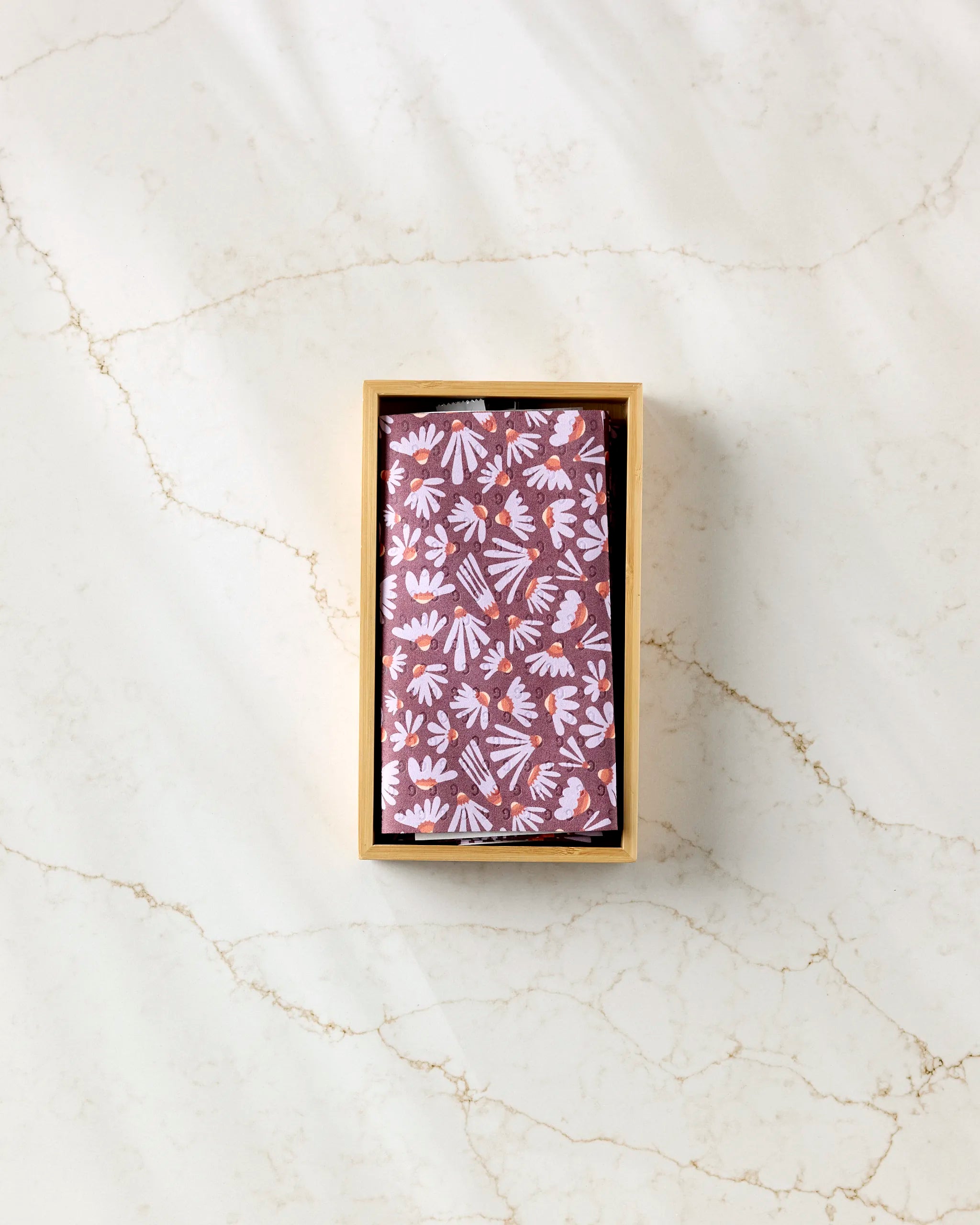 A rectangular wooden box from Elisabeth Wing contains Flower Pop, wrapped in brown paper with a white and lavender floral pattern, set on a light marble surface with subtle veining.