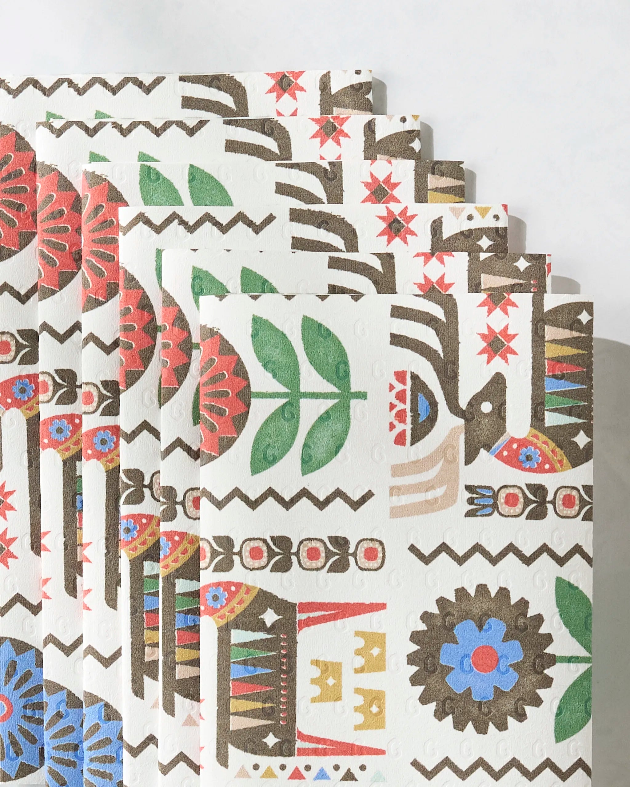Folk Deer by Geometry: eco-friendly paper towels featuring bold folk-style patterns—floral designs, geometric shapes, birds, leaves, and houses in vivid red, blue, green, brown, and yellow on a white background.