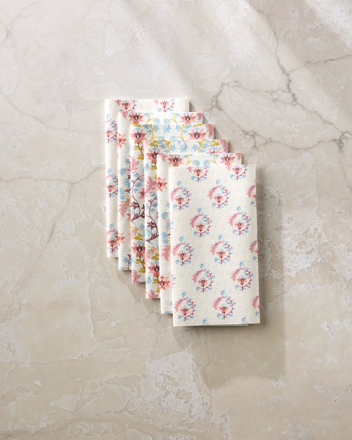 Five Folk Medallion cloth napkins by Maayan Alper-Swan, featuring assorted pastel floral patterns, are neatly stacked on a light beige marble surface, perfect as eco-friendly paper towel alternatives.