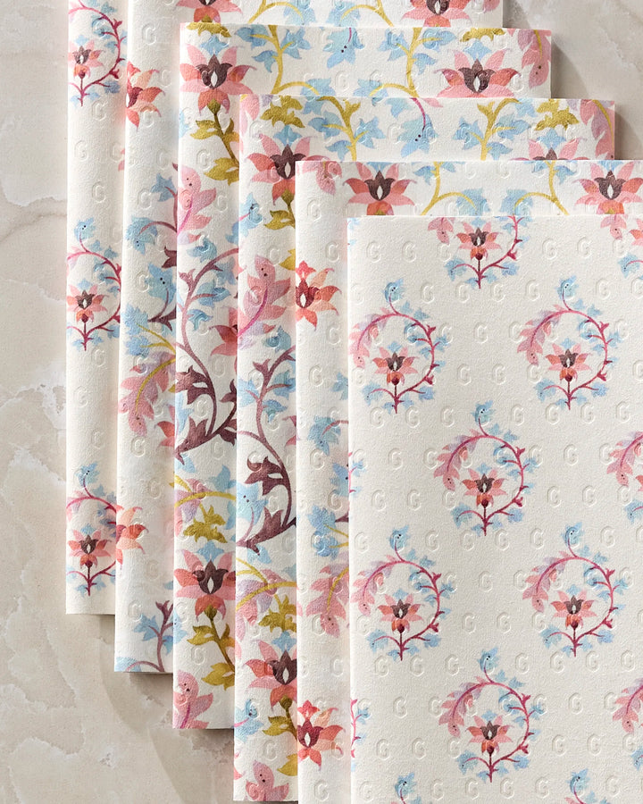 Five sheets of Maayan Alper-Swan Folk Medallion eco-friendly paper towels, featuring pastel pink, blue, yellow, and brown floral prints with an embossed G monogram, are fanned out on a marbled surface.